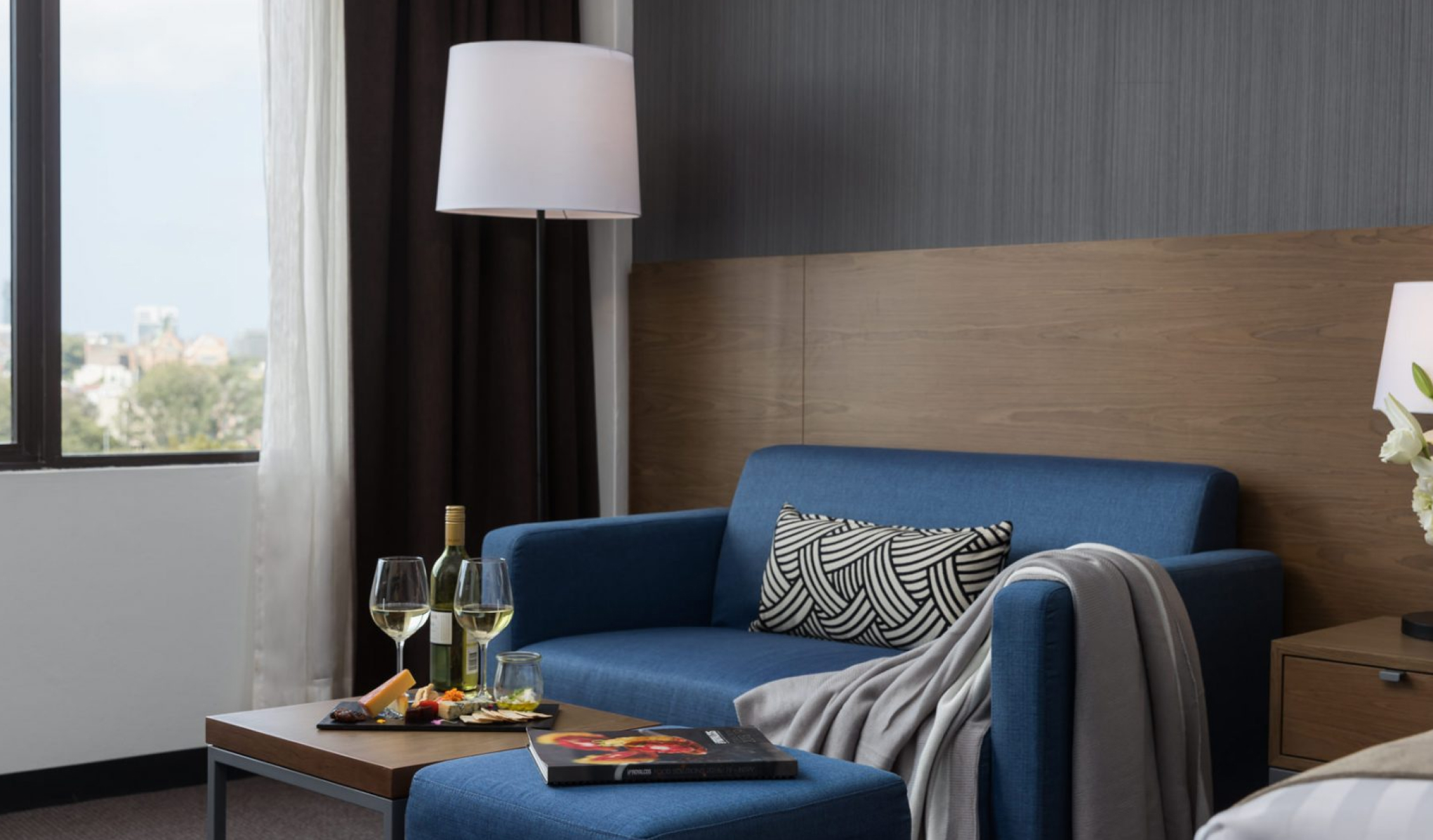 Blue sofa and ottoman with patterned cushions and a draped blanket, beside a floor lamp, coffee table, and window.