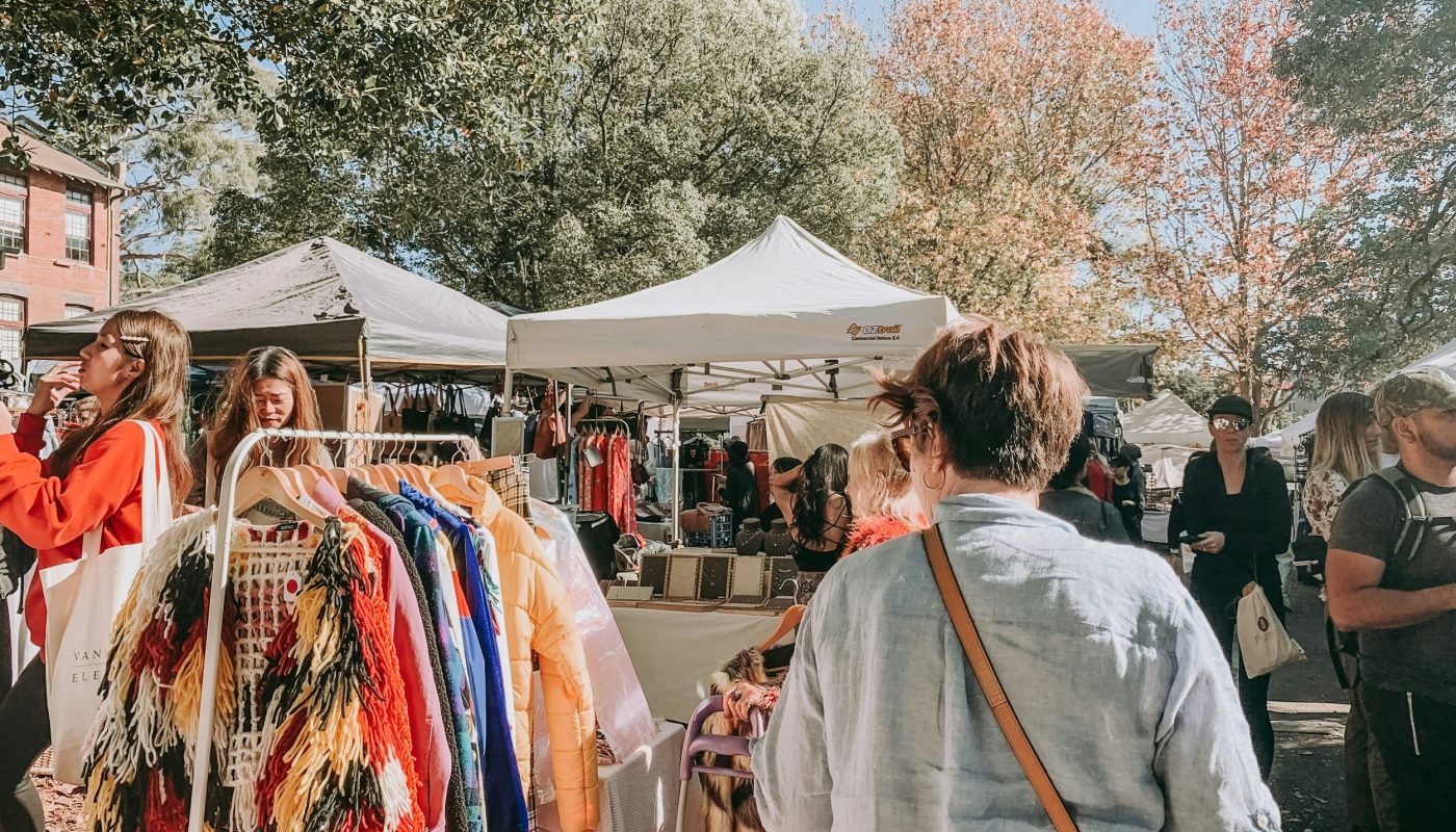 People browse clothing racks at an outdoor market beneath canopy tents, with leafy trees in the background.