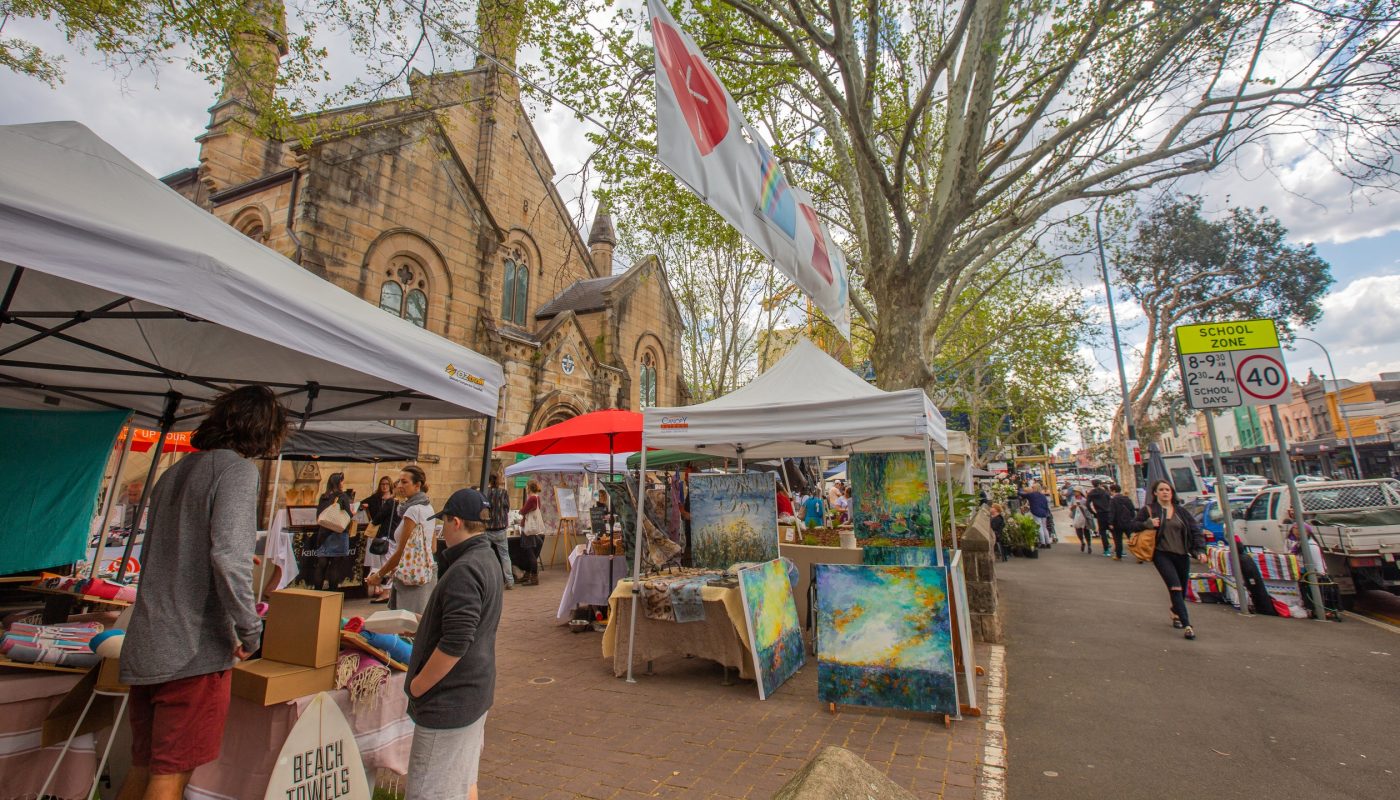 Outdoor market stalls line a tree-shaded street beside a stone church, with vendors and shoppers.