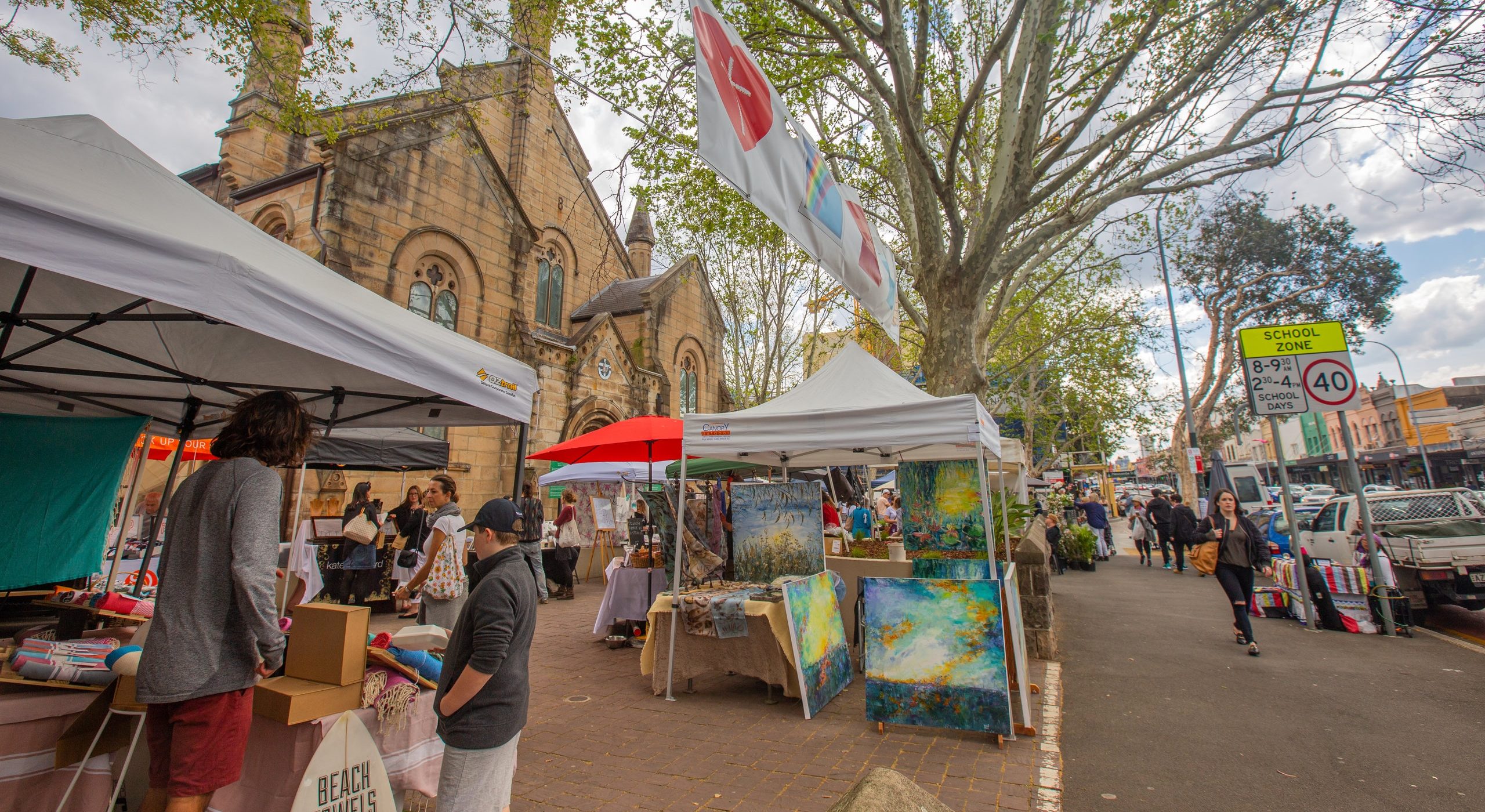 Outdoor market stalls line a tree-shaded street beside a stone church, with vendors and shoppers.