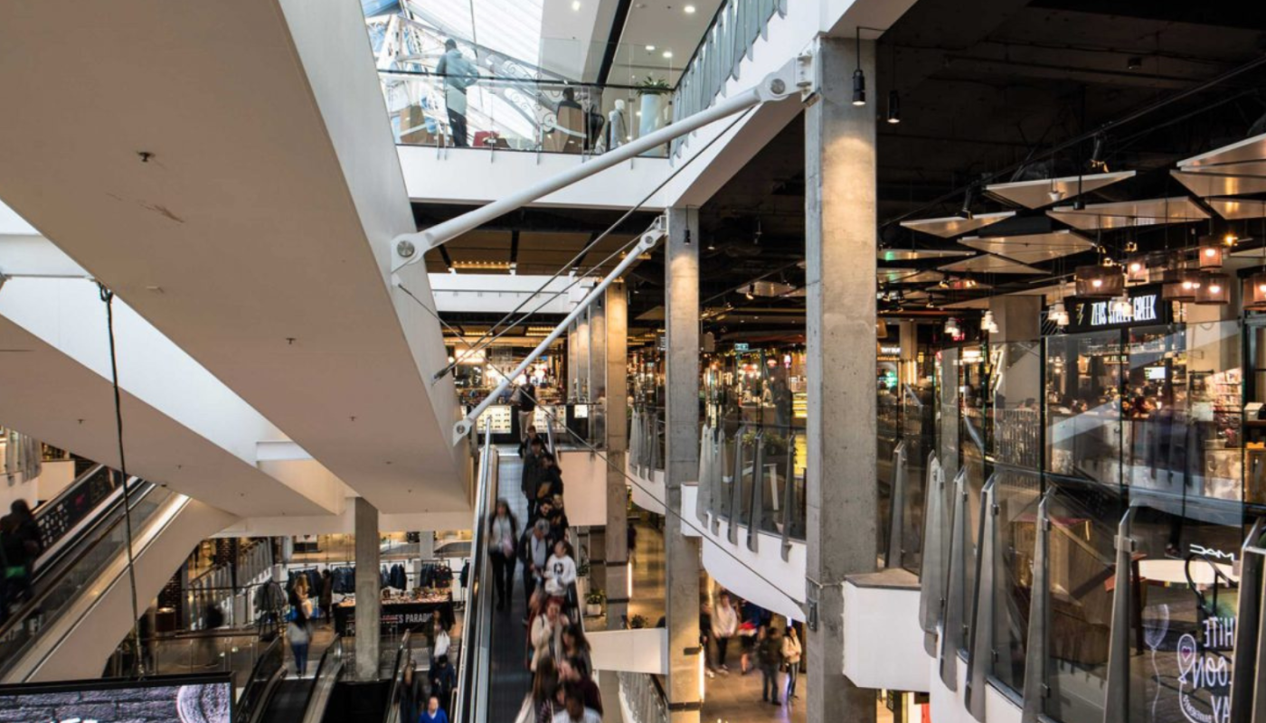 Multi-level shopping mall atrium with escalators, glass railings, storefronts, and people walking below.