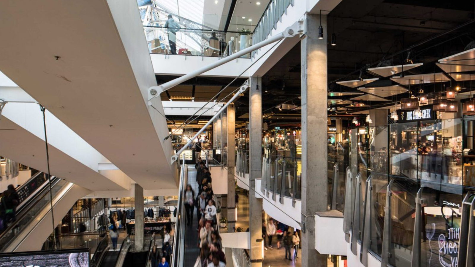 Multi-level shopping mall atrium with escalators, glass railings, storefronts, and people walking below.
