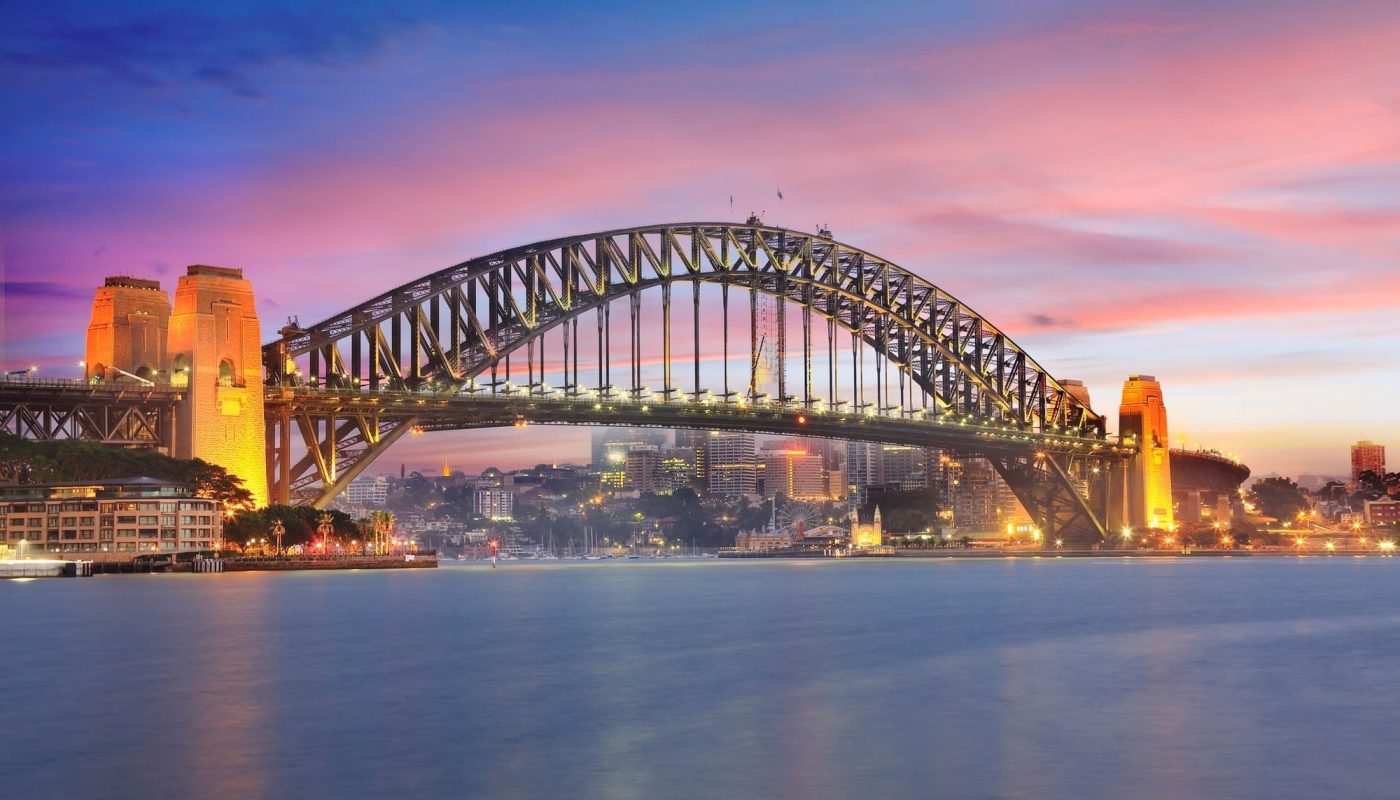Sydney Harbour Bridge spans the harbor at sunset, with illuminated buildings along the shore and calm water in the foreground.