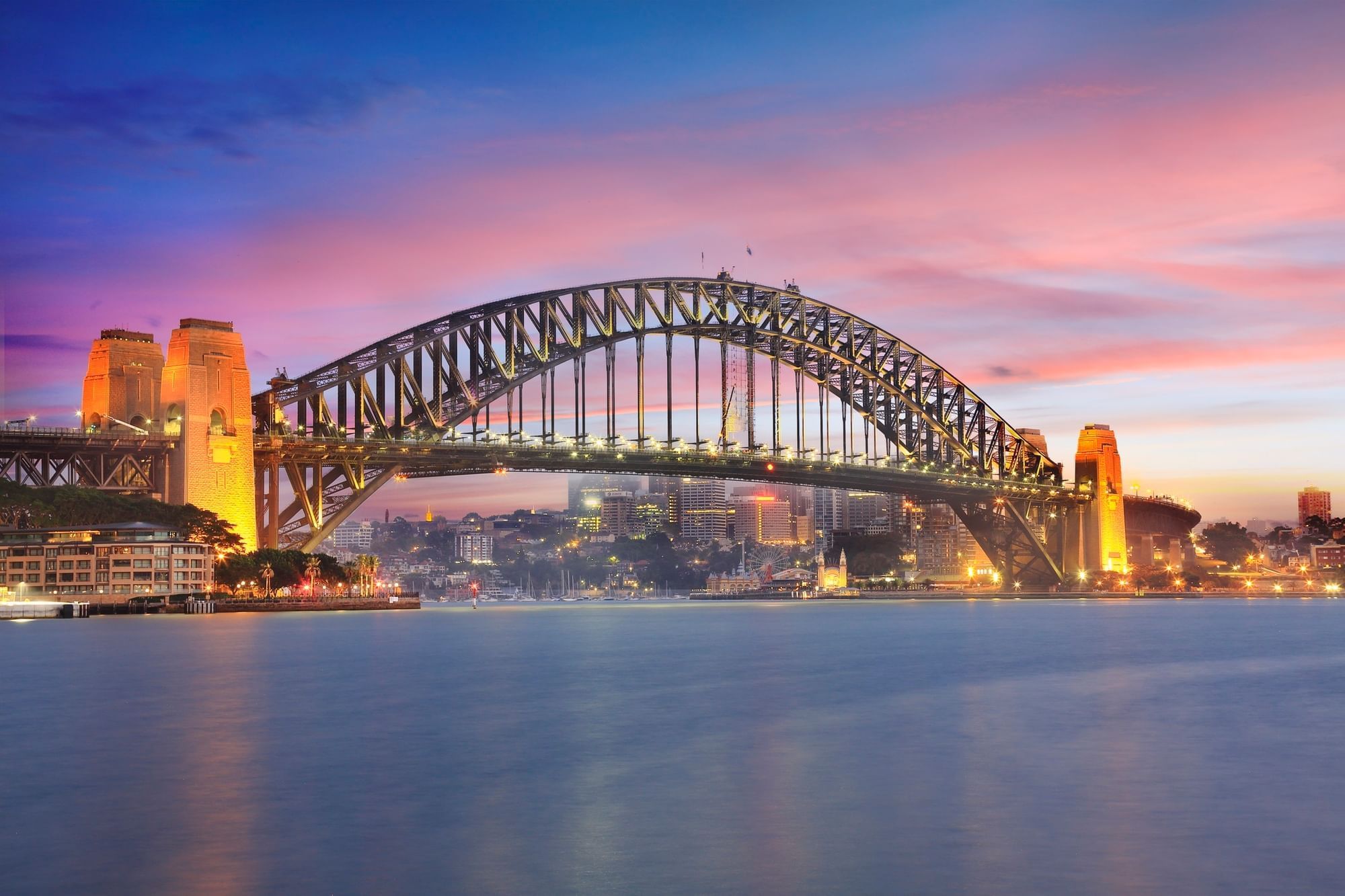 Sydney Harbour Bridge spans the harbor at sunset, with illuminated buildings along the shore and calm water in the foreground.
