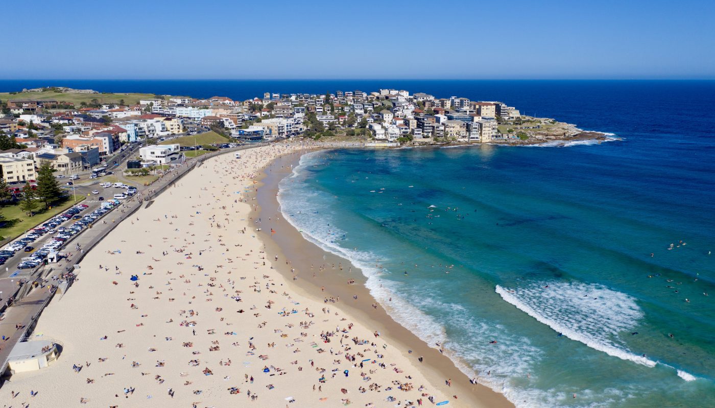 Aerial view of a crescent-shaped beach with people, turquoise surf, and a coastal town along the shore.