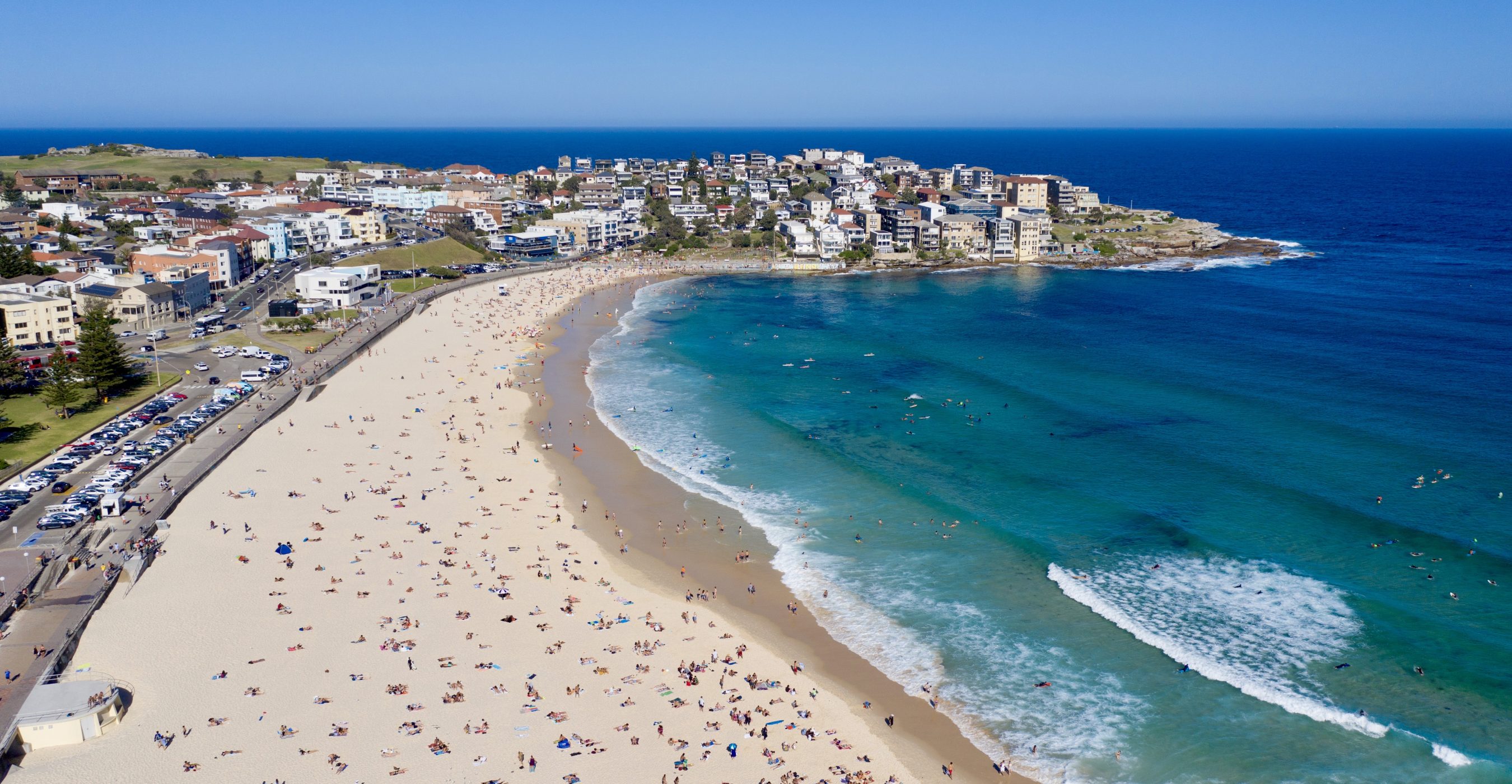 Aerial view of a crescent-shaped beach with people, turquoise surf, and a coastal town along the shore.