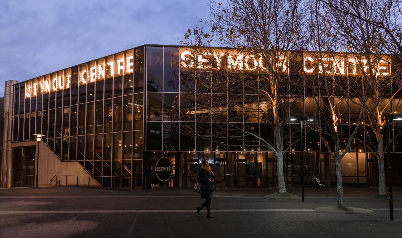 Glass-fronted building with illuminated signage at dusk, leafless trees along the sidewalk, and a pedestrian crossing in front.