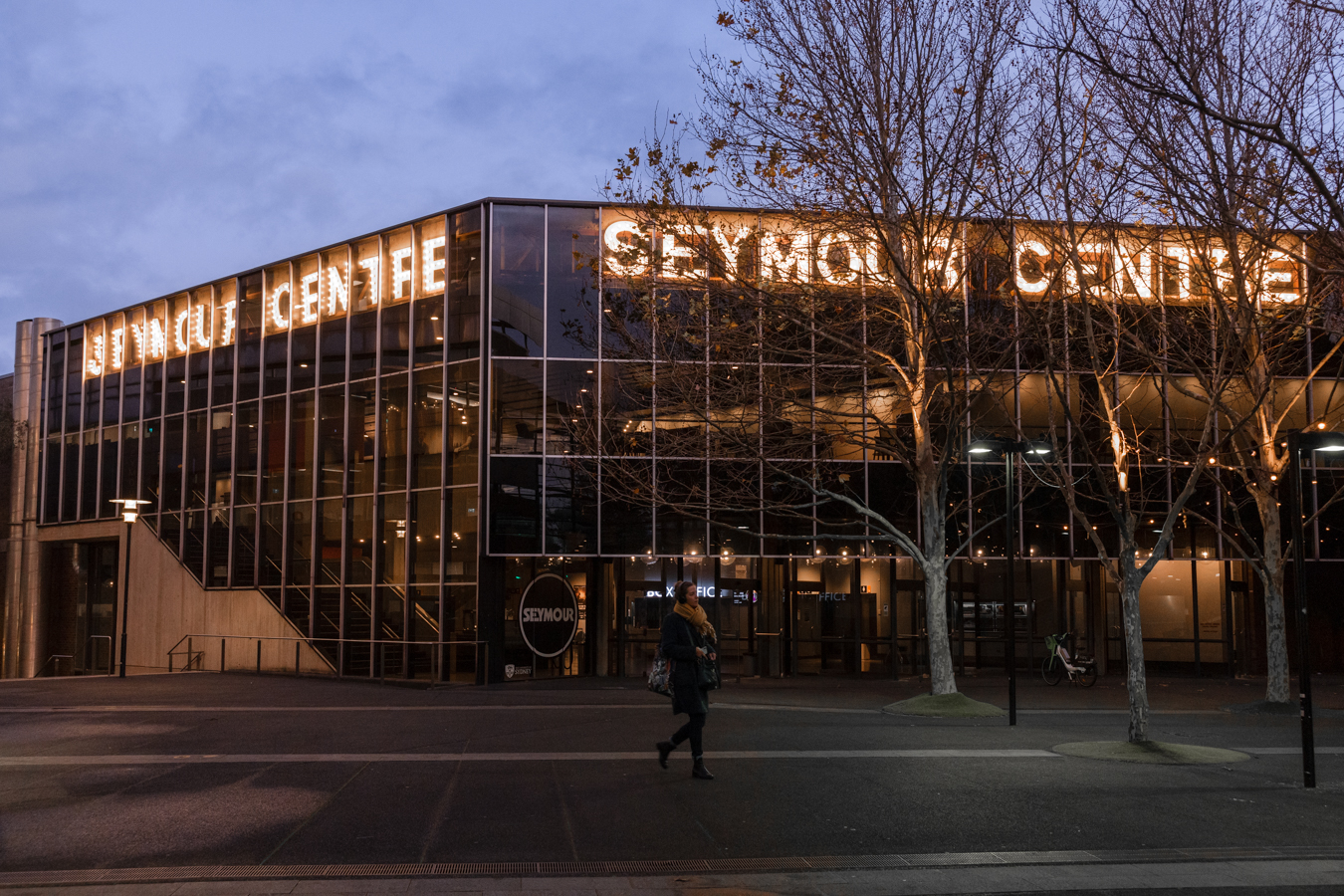 Glass-fronted building with illuminated signage at dusk, leafless trees along the sidewalk, and a pedestrian crossing in front.
