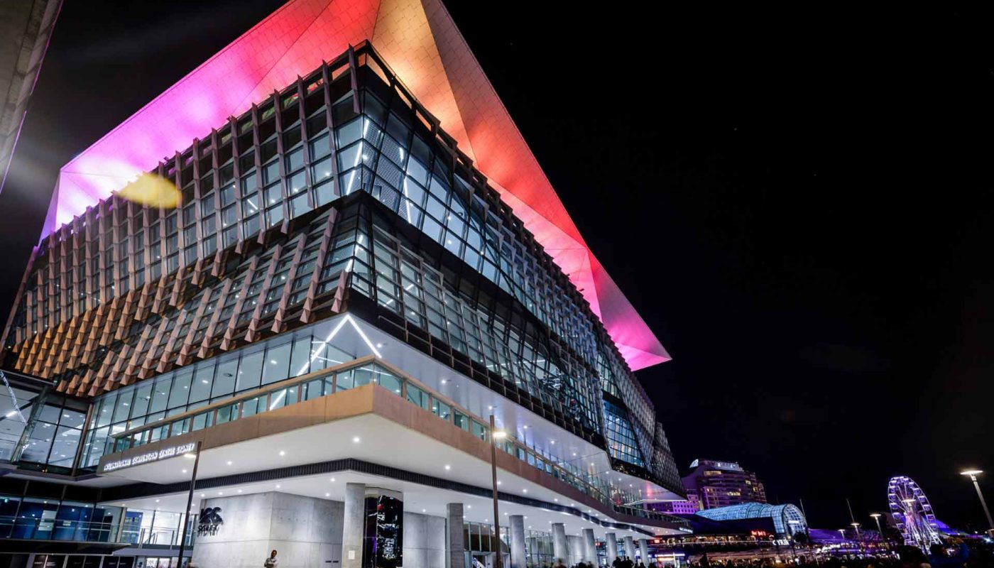 Modern glass building at night with a sharply angled roof glowing pink and orange.