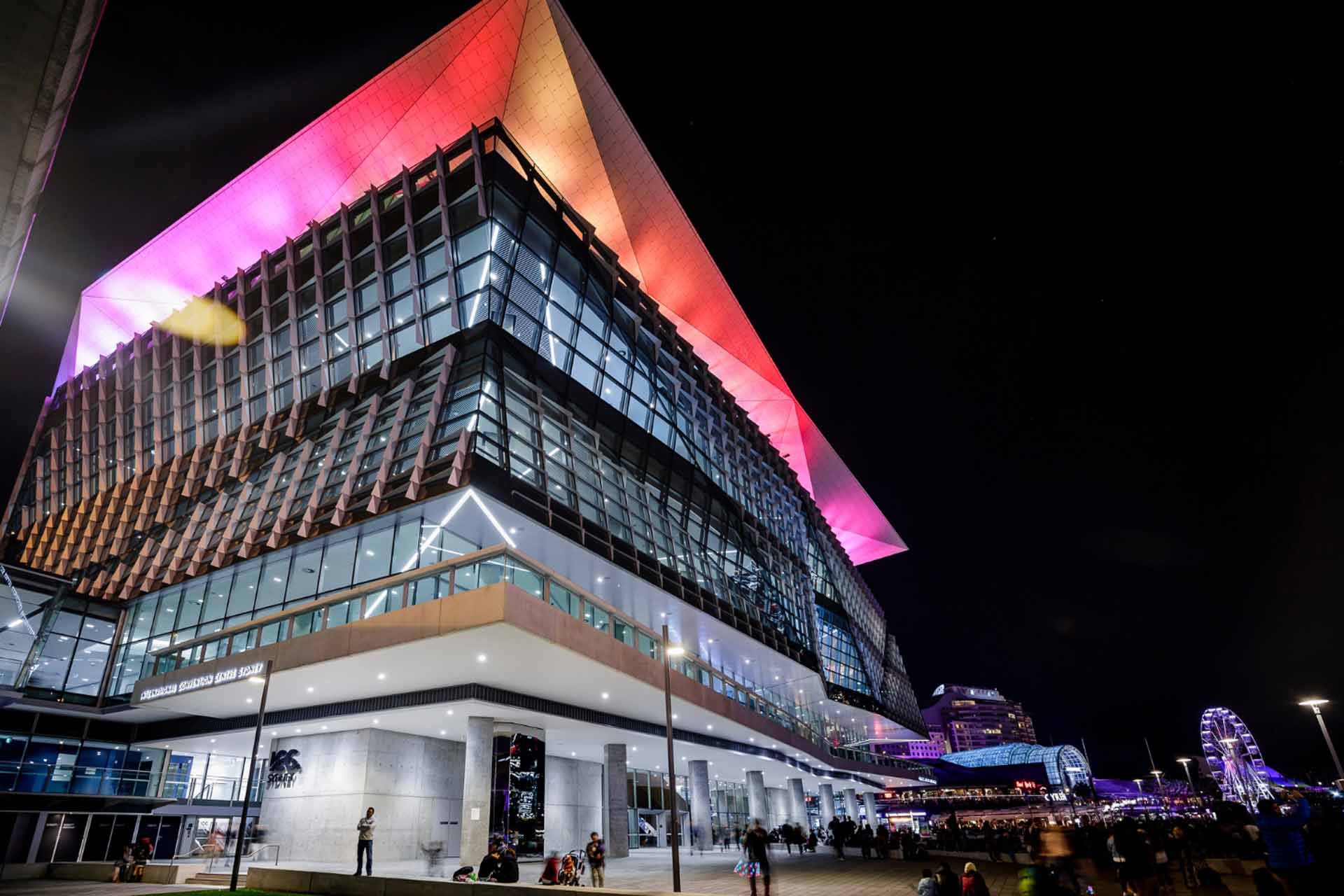 Modern glass building at night with a sharply angled roof glowing pink and orange.