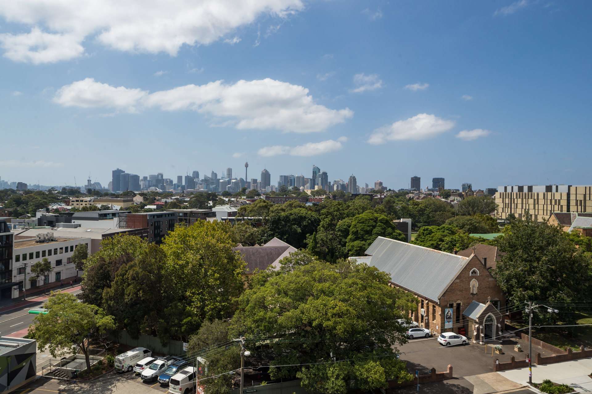 A city skyline rises beyond leafy residential buildings beneath a blue sky with scattered clouds.
