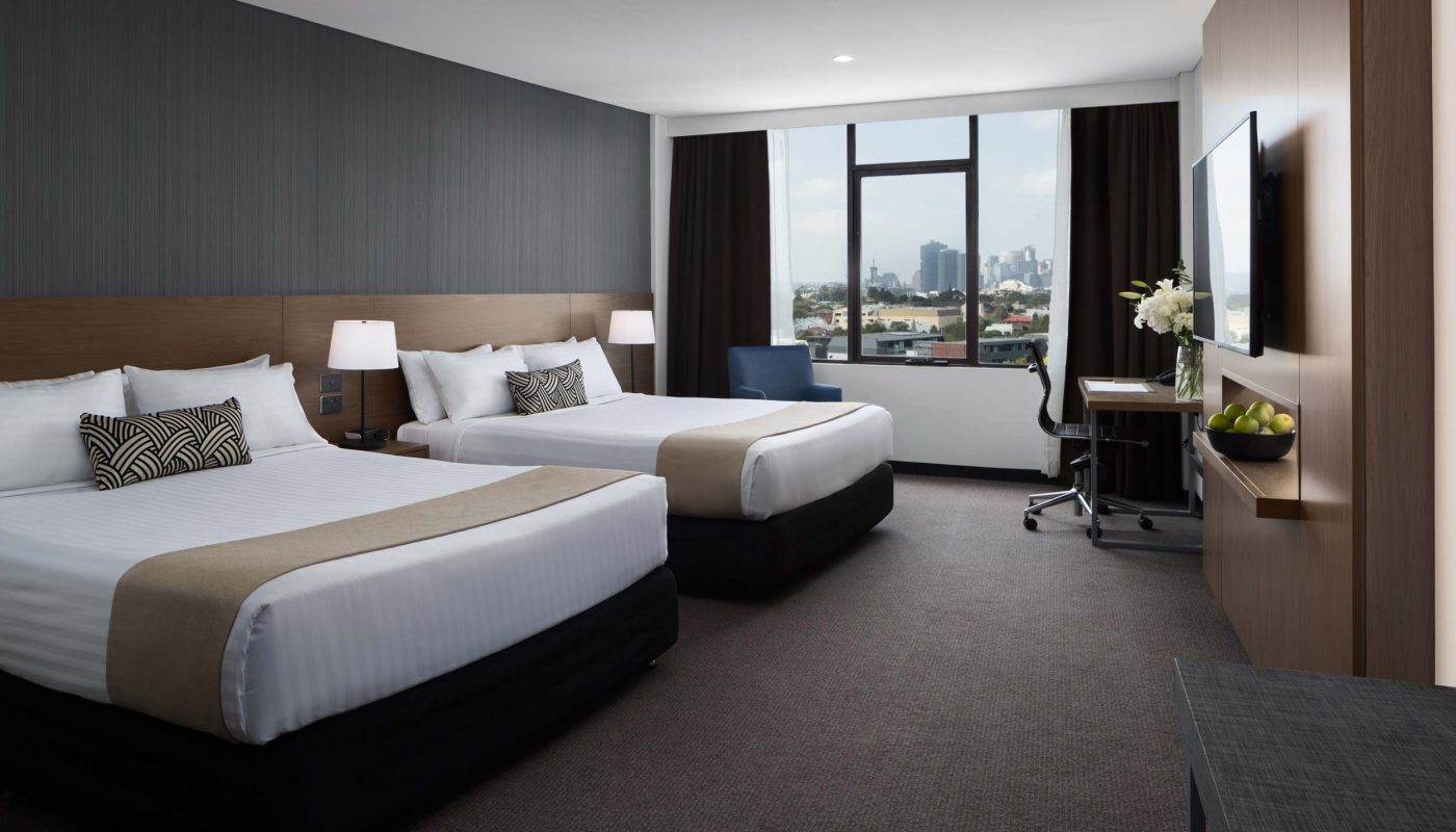 Modern hotel room with two beds, a large window overlooking the city, and a desk beside a wall-mounted TV.