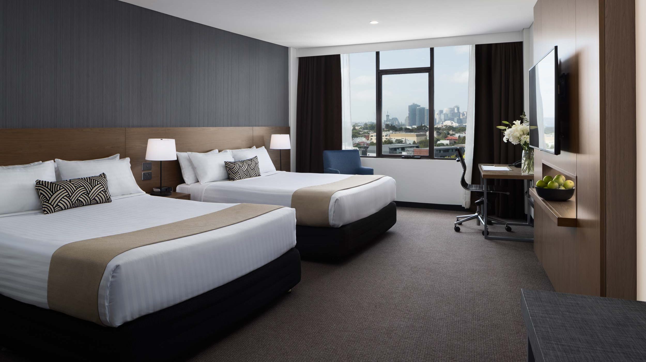 Modern hotel room with two beds, a large window overlooking the city, and a desk beside a wall-mounted TV.