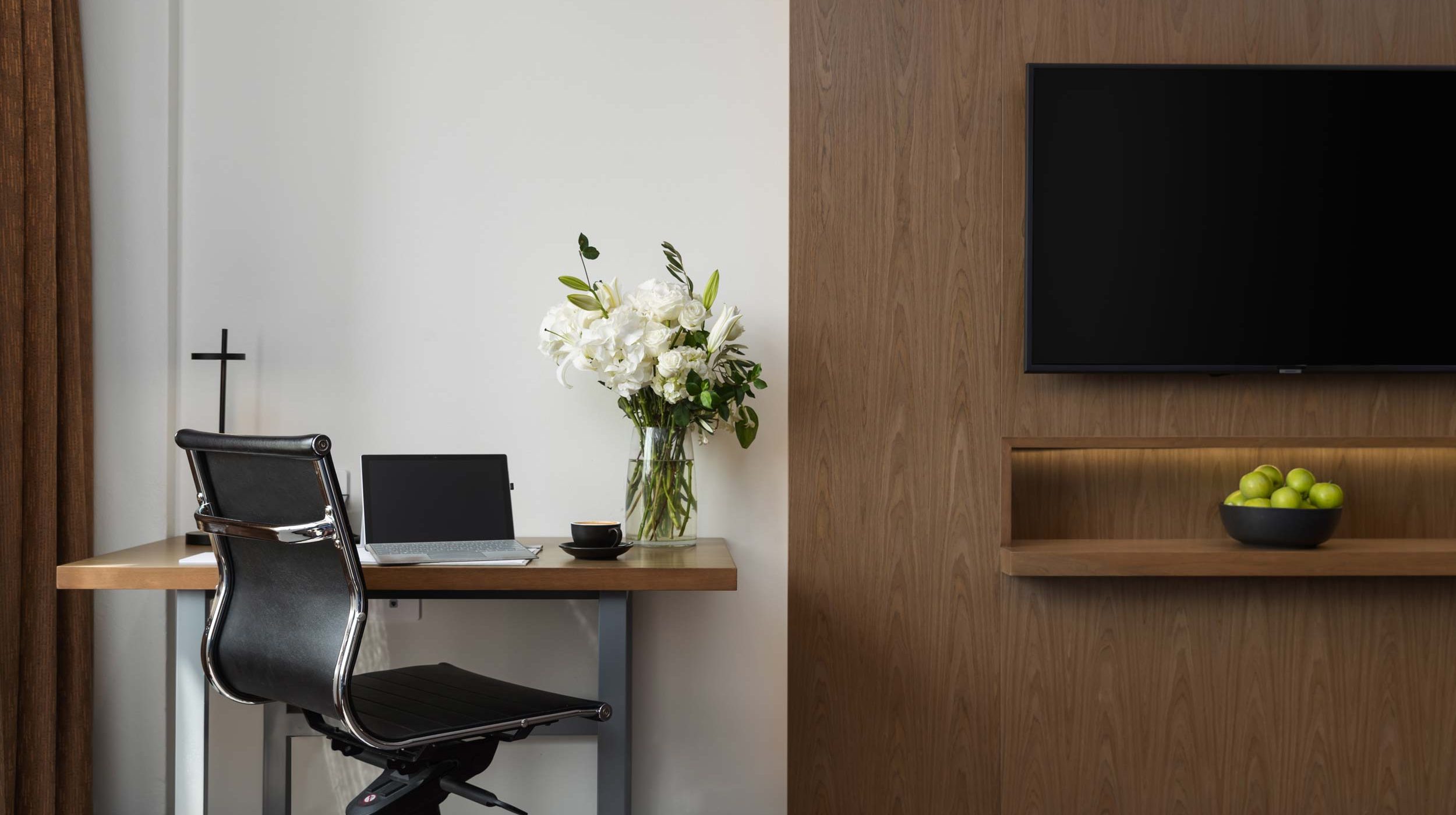Desk and black chair beside a wall-mounted TV, with white flowers and a bowl of green apples.