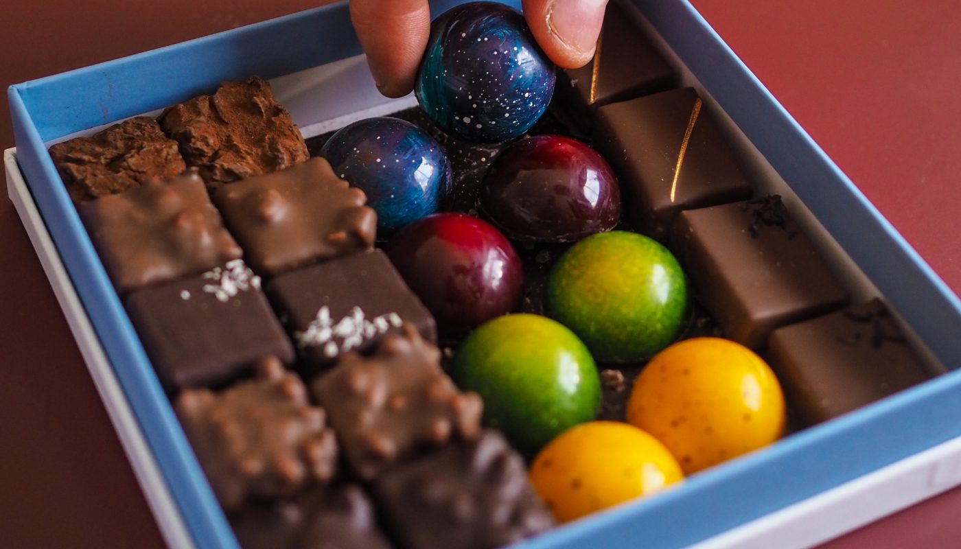 A hand picks up a blue round chocolate from a blue box filled with assorted chocolates.