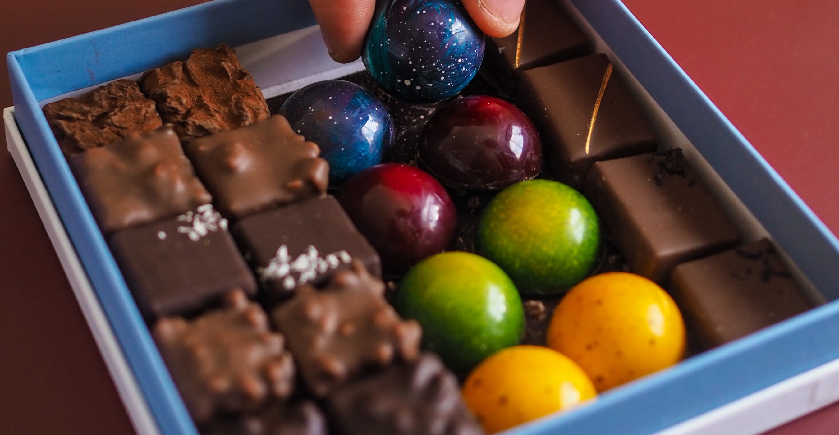 A hand picks up a blue round chocolate from a blue box filled with assorted chocolates.
