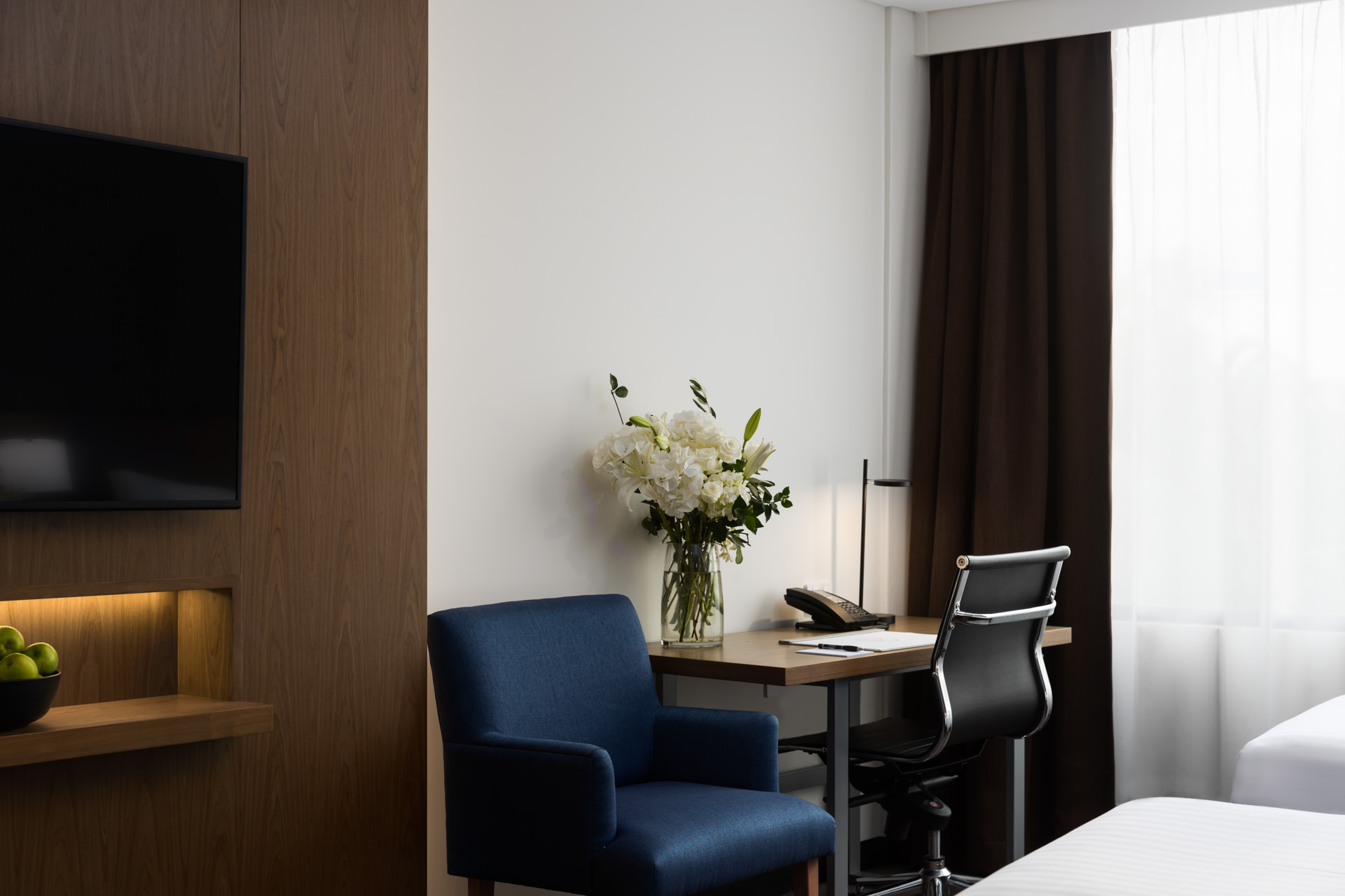 Modern hotel room with a wall-mounted TV, blue armchair, desk and chair, flowers, and a bed beside curtained windows.