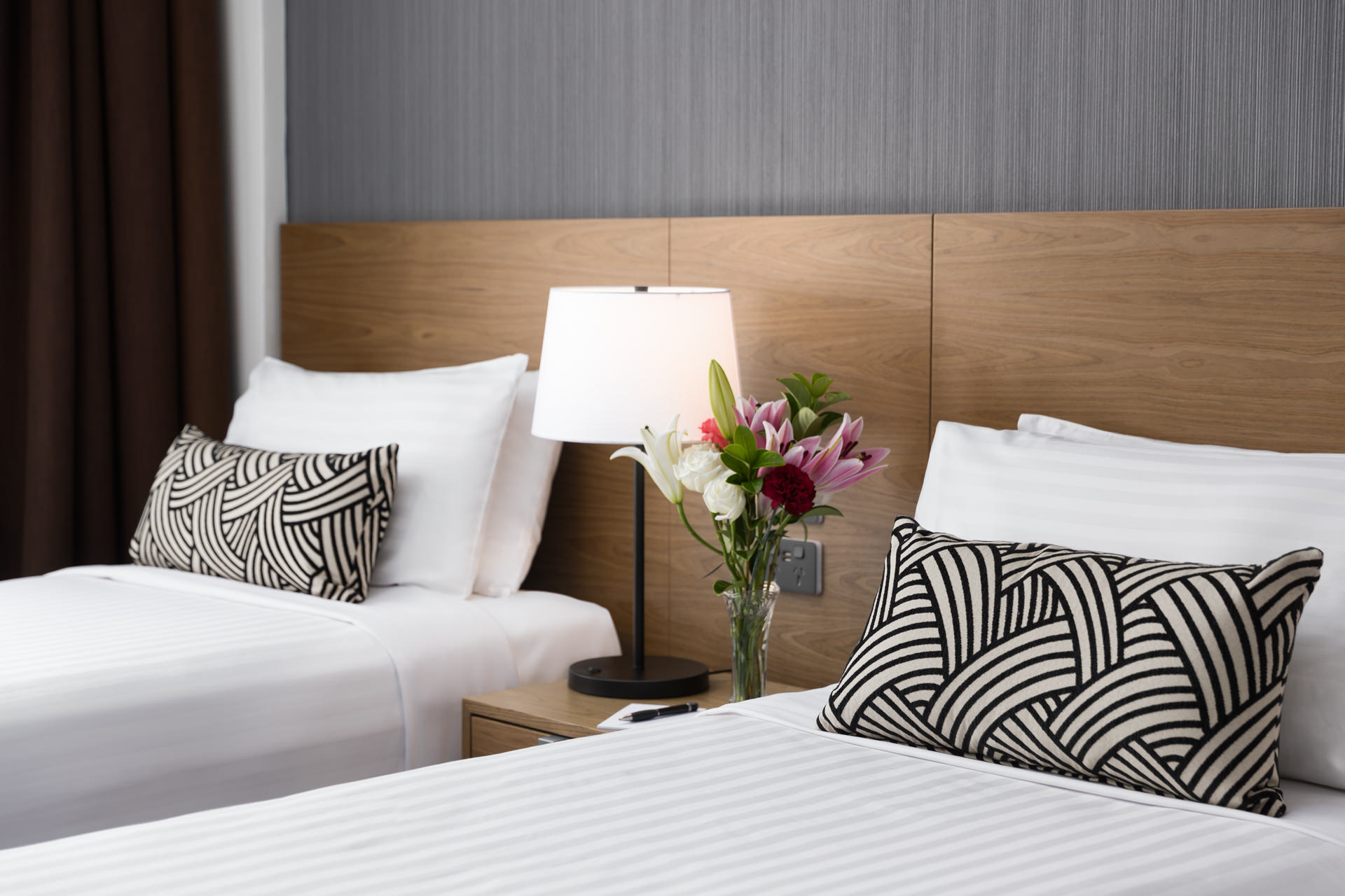 Two neatly made beds with patterned pillows, a bedside lamp, and a vase of flowers between them.