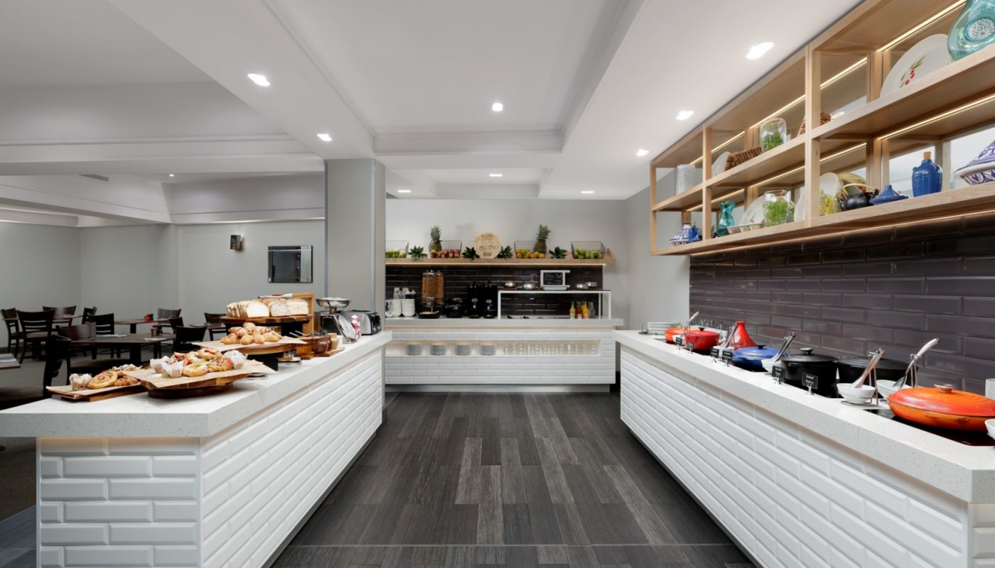 Bright commercial kitchen with white counters, food trays, open shelving, and a dark tiled floor.