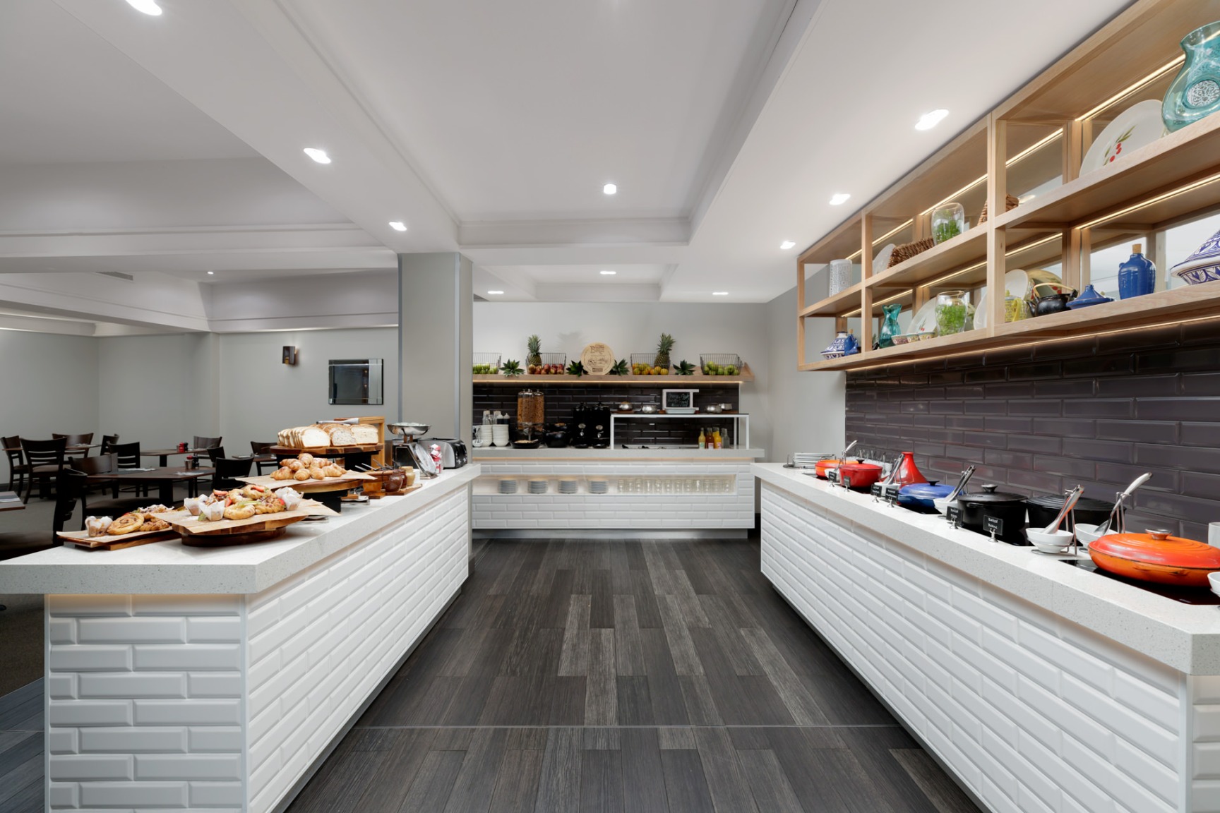 Bright commercial kitchen with white counters, food trays, open shelving, and a dark tiled floor.