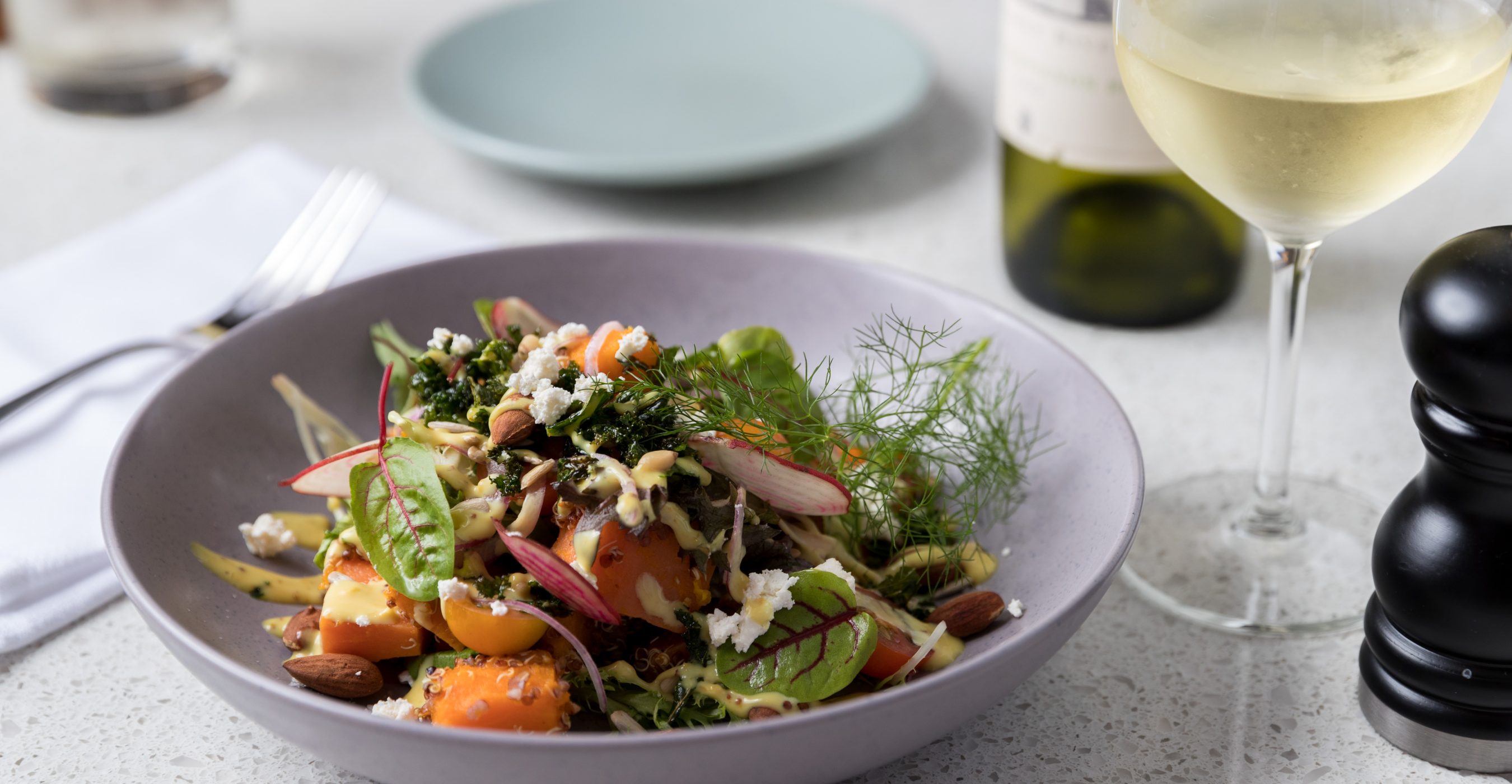A colorful salad with leafy greens, sliced vegetables, nuts, crumbled cheese, and creamy dressing in a gray bowl.