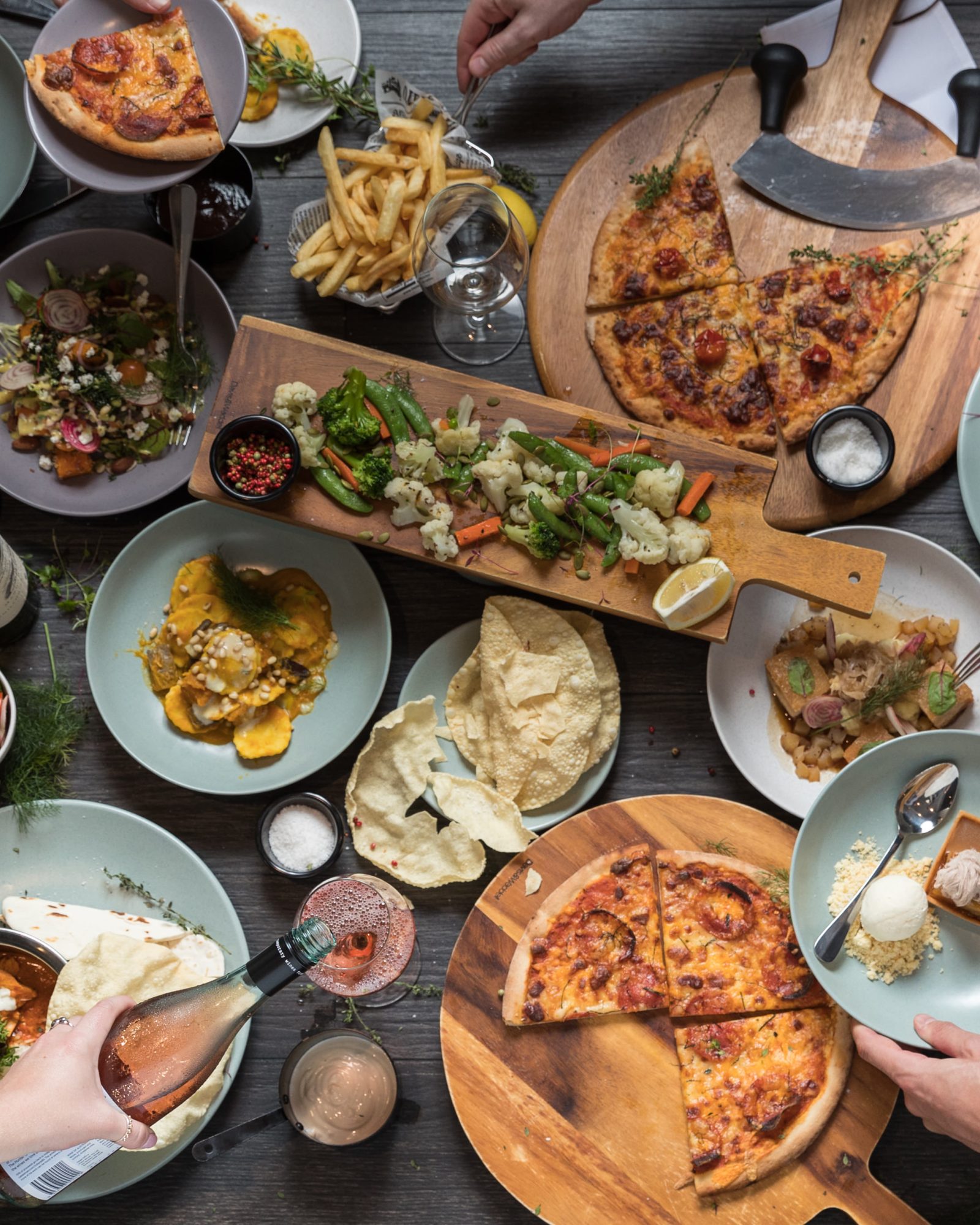 Overhead view of a crowded table with pizzas, salads, and shared dishes as diners reach for food.