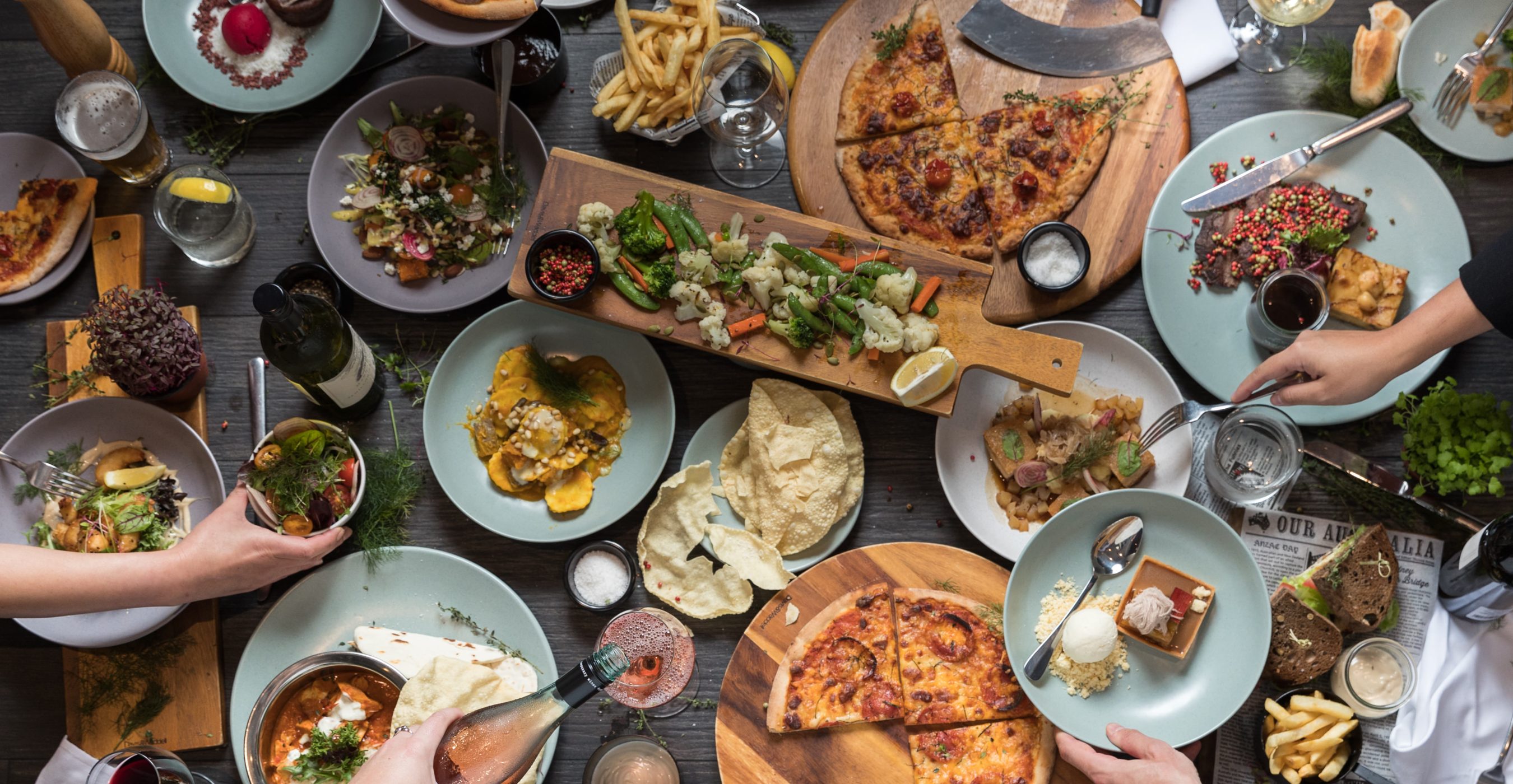 Overhead view of a crowded table with pizzas, salads, and shared dishes as diners reach for food.