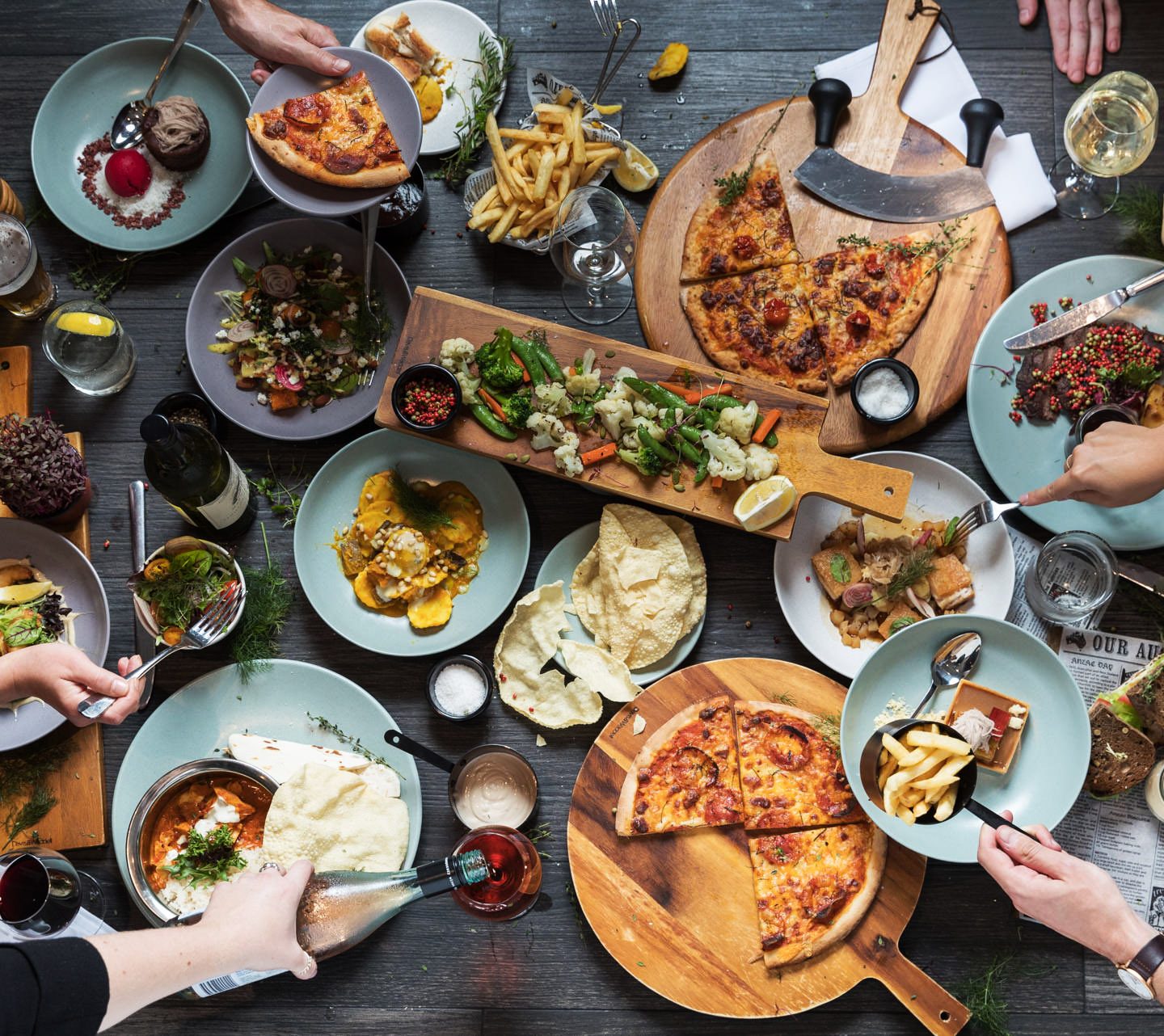 Overhead view of hands sharing pizzas, salads, bread, and other dishes around a crowded dining table.