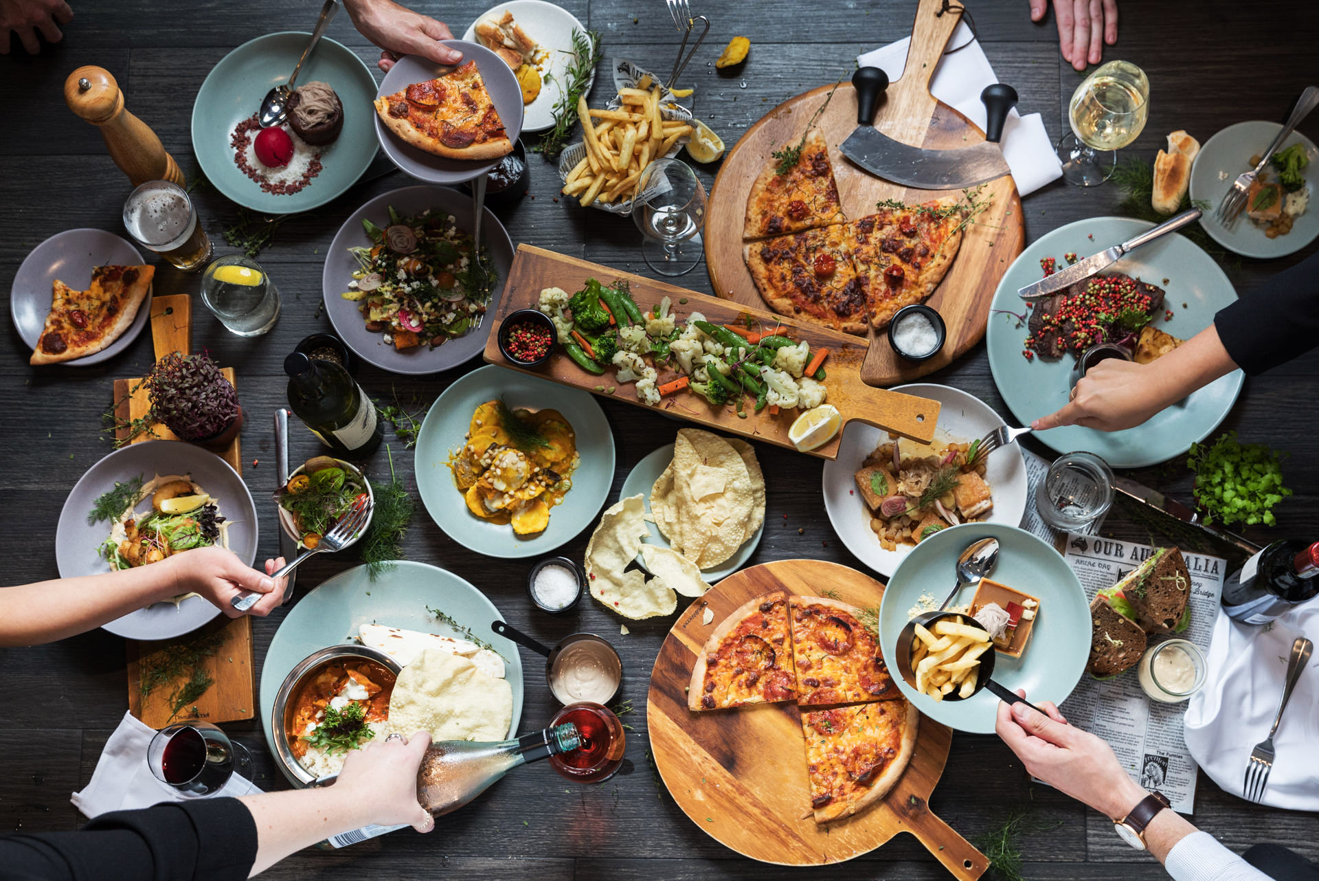 Overhead view of hands sharing pizzas, salads, bread, and other dishes around a crowded dining table.