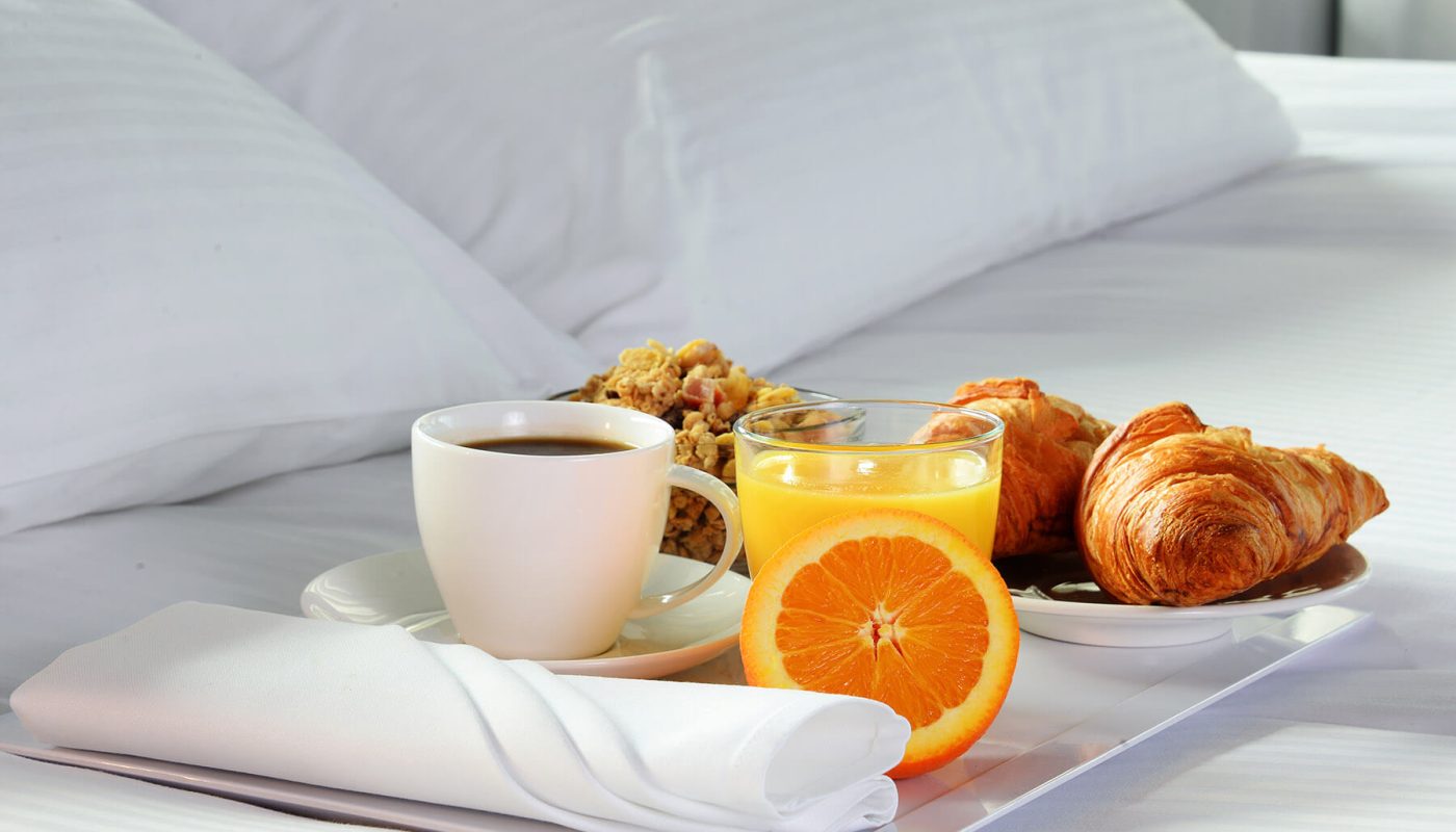 Breakfast on a tray atop white bedding, with coffee, orange juice, an orange half, and croissants.