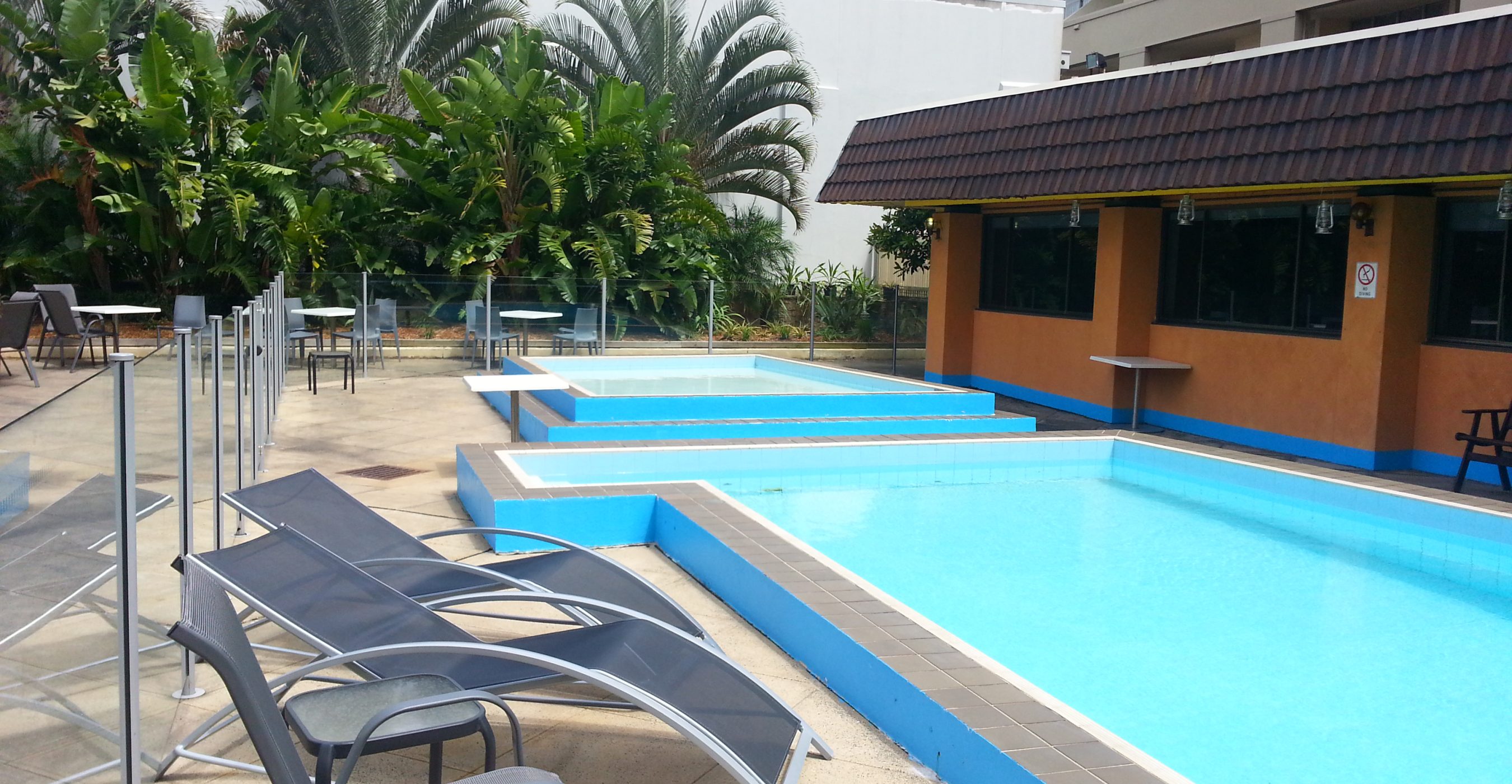 A bright blue outdoor pool beside lounge chairs, palm trees, and a low building.