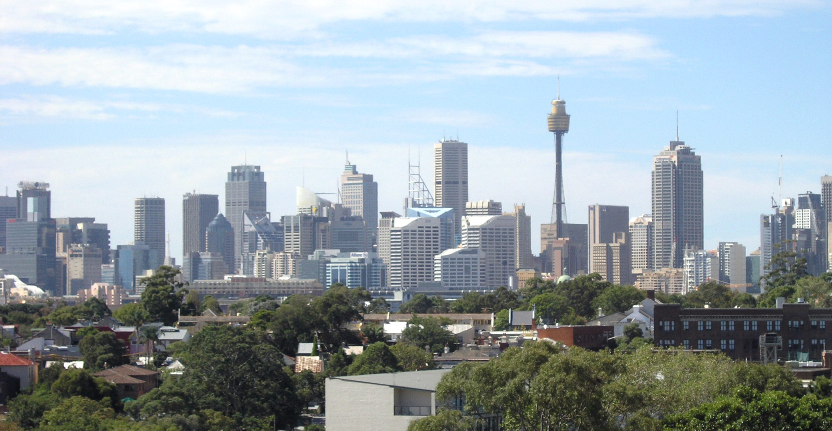 A city skyline rises behind leafy residential rooftops under a blue, partly cloudy sky.