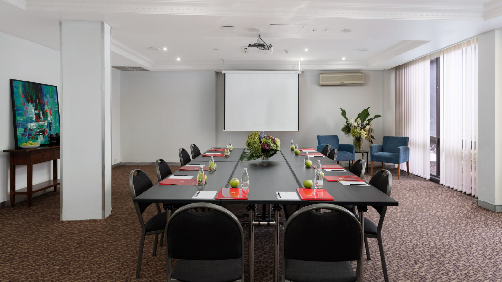 A conference room with a long table and chairs, a wall screen, large windows, and potted plants.