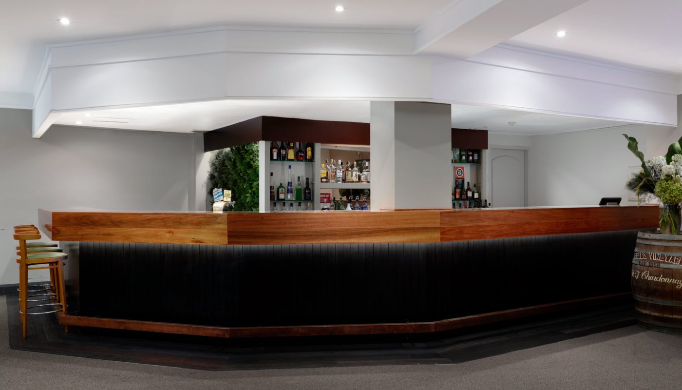 A long dark bar with wood trim sits beneath liquor shelves in a modern interior.