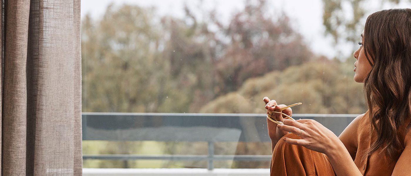 Woman seated beside a window, holding eyeglasses and looking out toward trees.