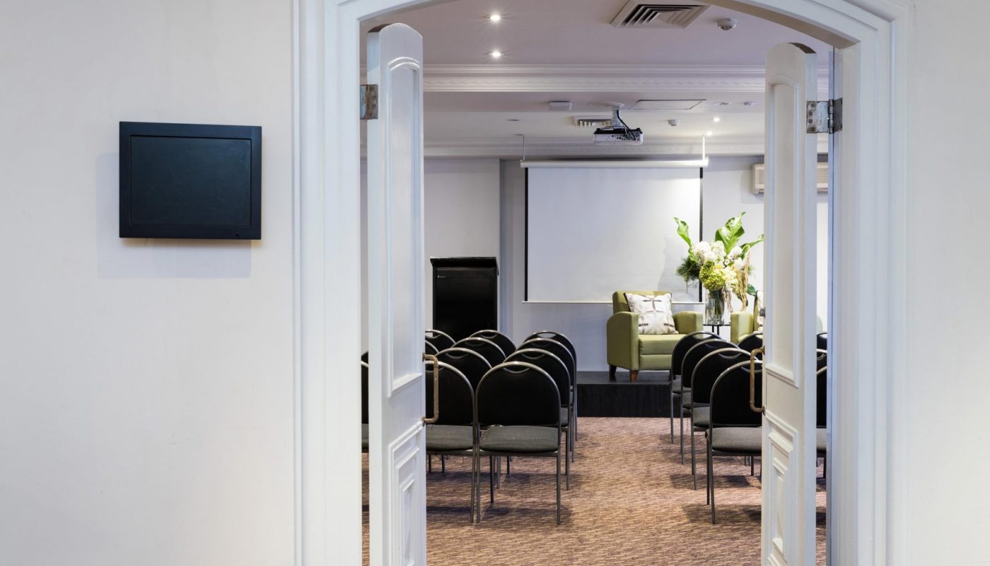 A meeting room viewed through open double doors, with rows of chairs facing a projector screen and green armchair.