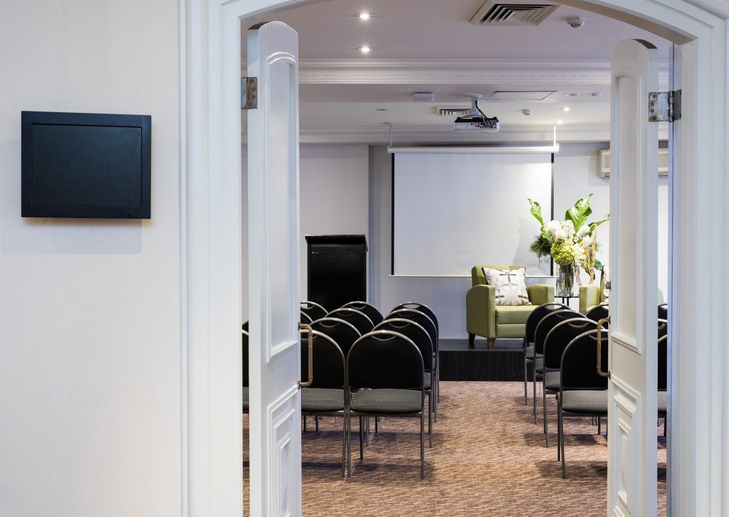 A meeting room viewed through open double doors, with rows of chairs facing a projector screen and green armchair.