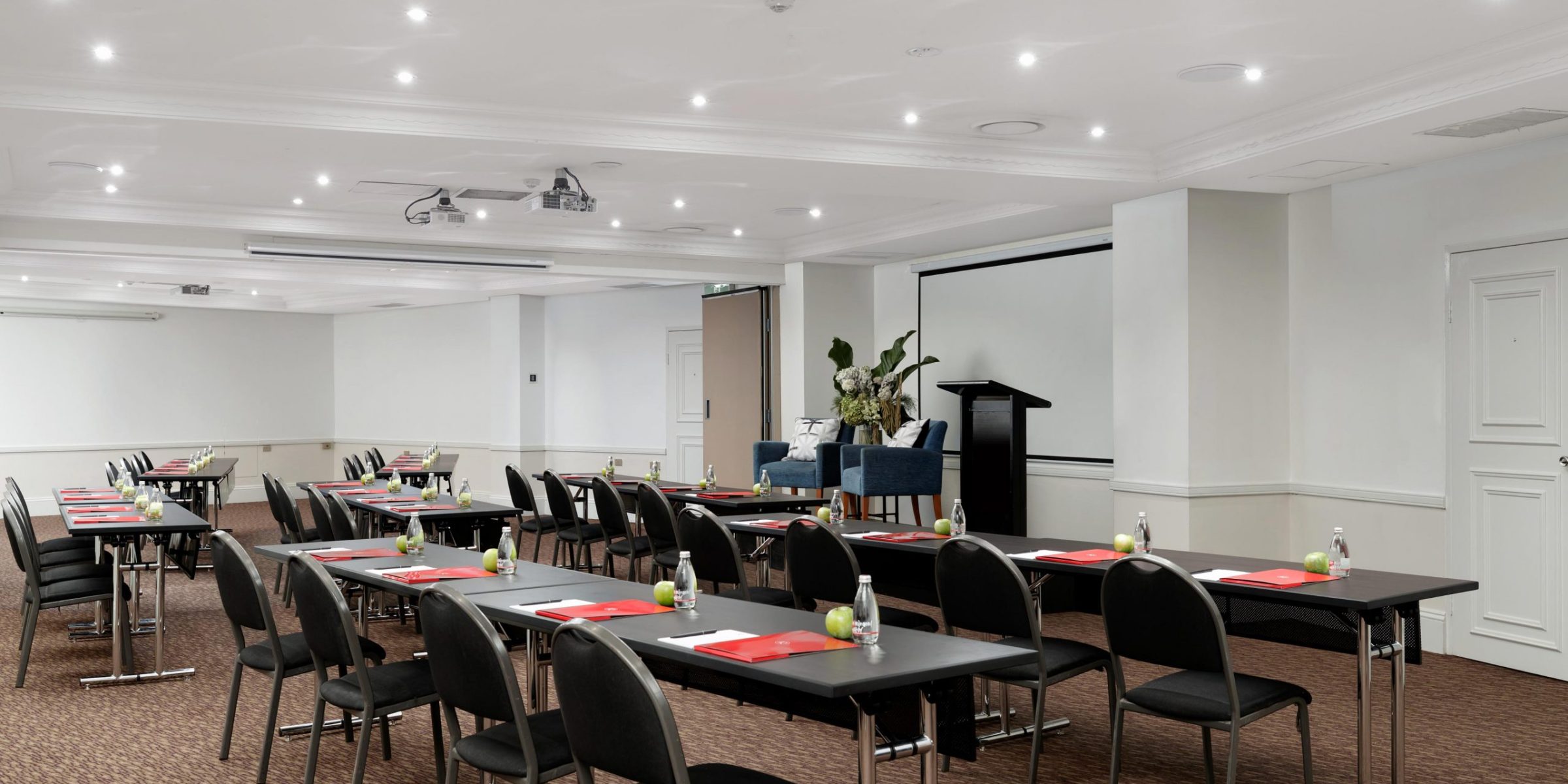 An empty conference room with rows of tables and chairs set with red folders, water bottles, and apples, facing a lectern and projection screen.