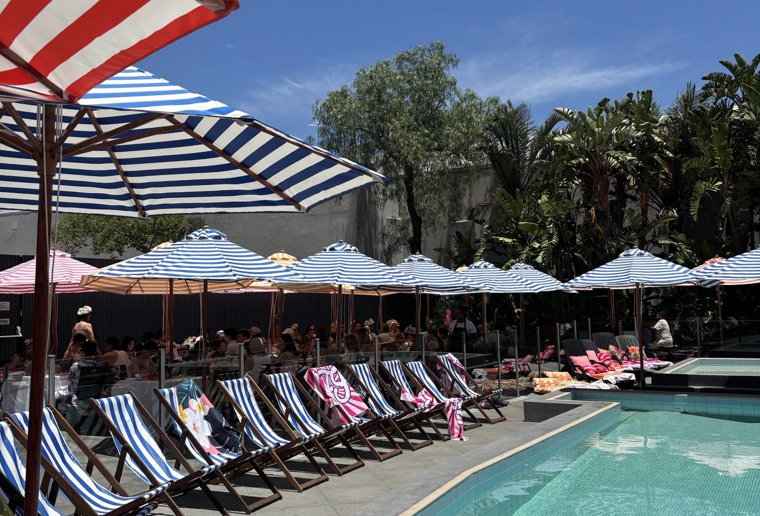 A sunny poolside terrace with striped umbrellas, lounge chairs, seated diners, and tropical plants.