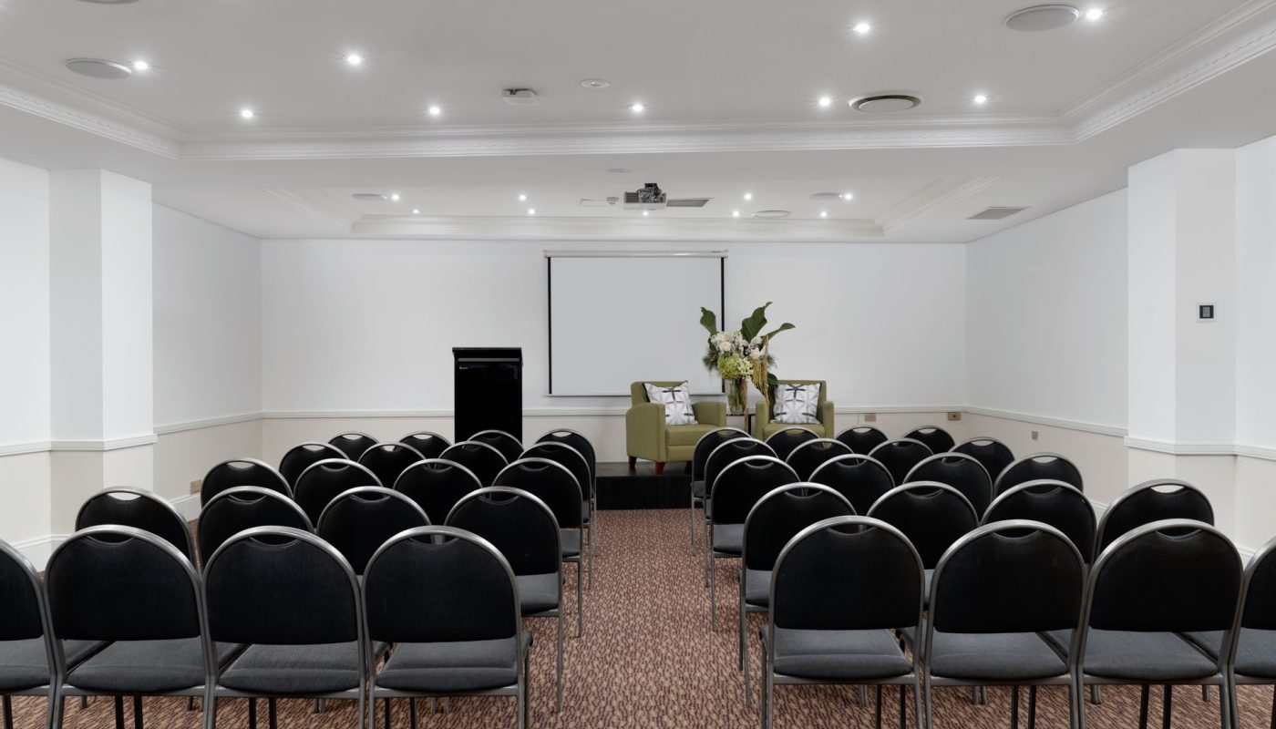 Empty conference room with rows of black chairs facing a stage with two armchairs, a lectern, and a projection screen.