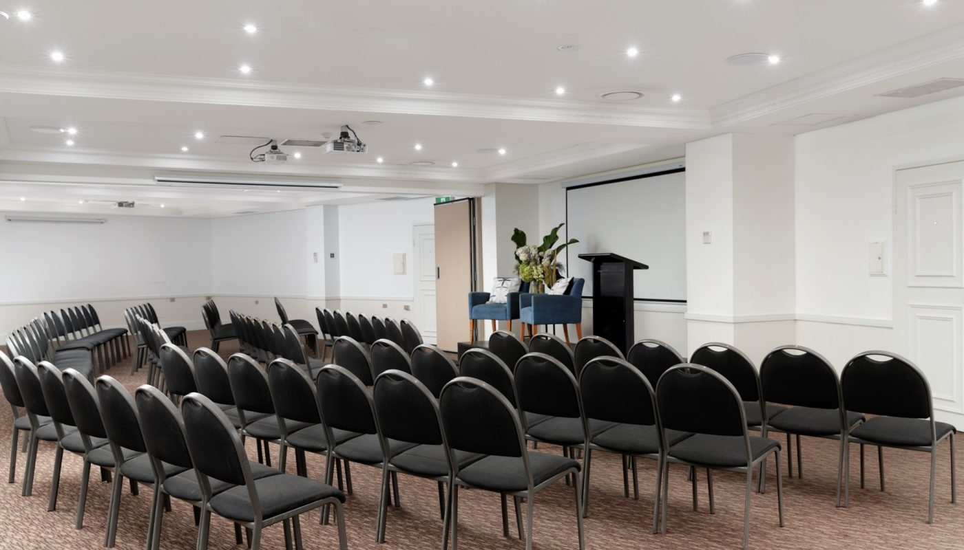 Rows of black chairs face a lectern and projection screen in a bright, empty conference room.