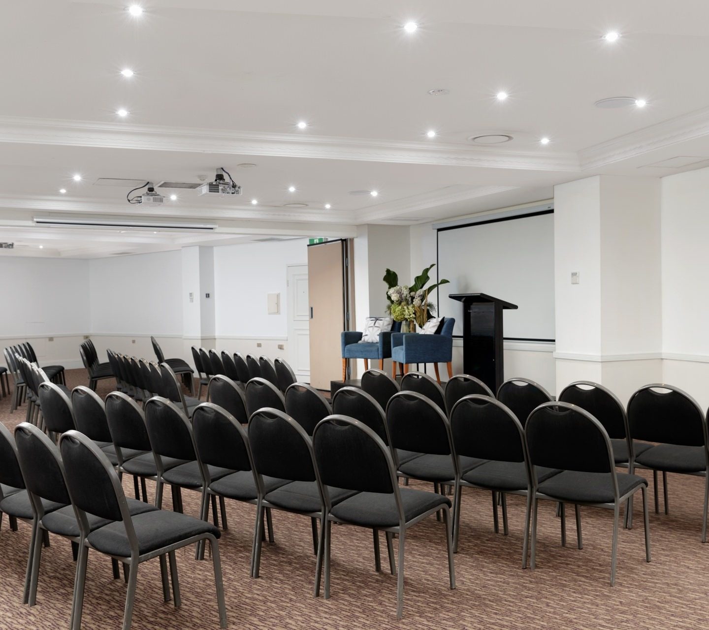 Rows of black chairs face a lectern and projection screen in a bright, empty conference room.