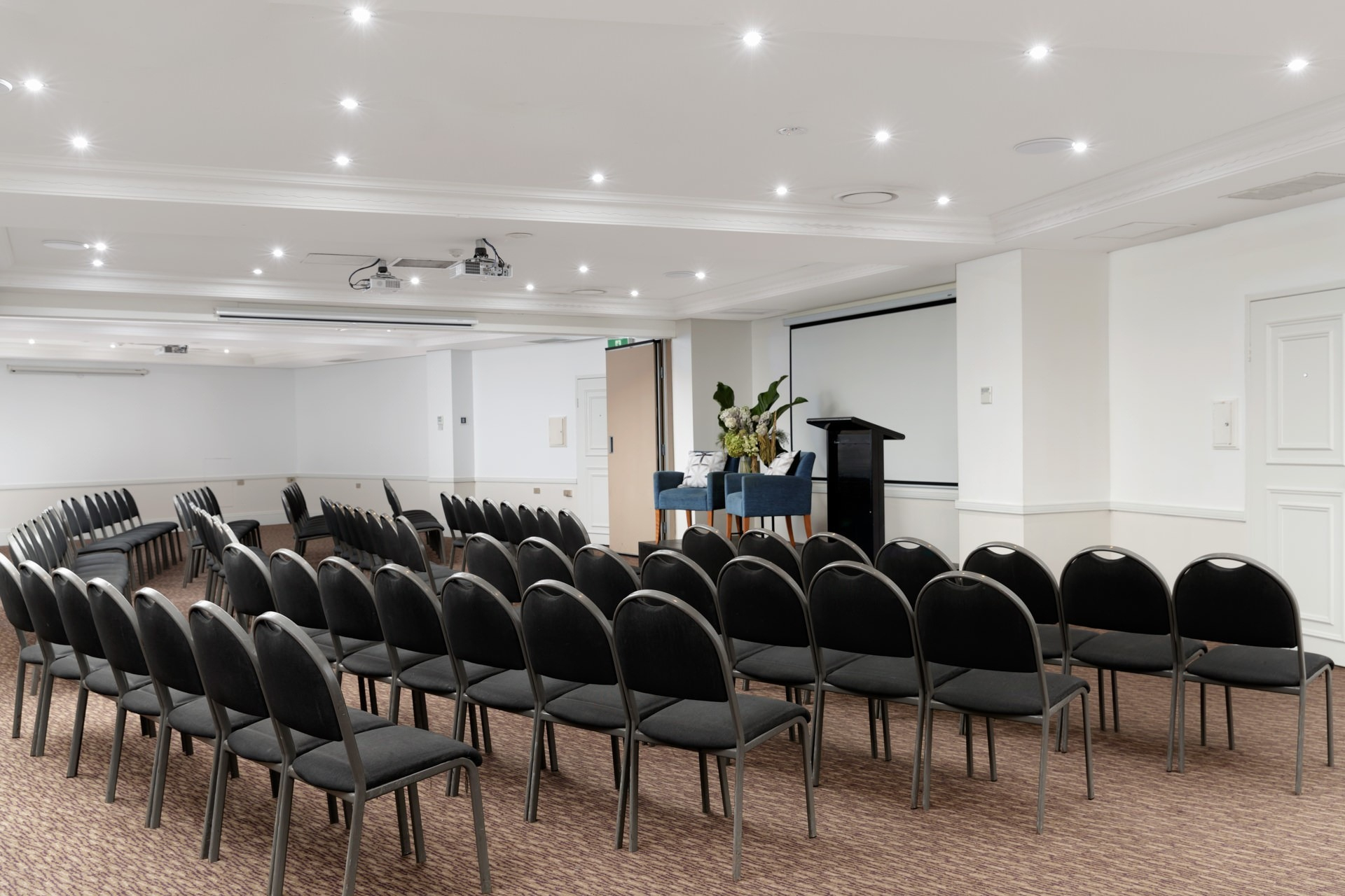 Rows of black chairs face a lectern and projection screen in a bright, empty conference room.