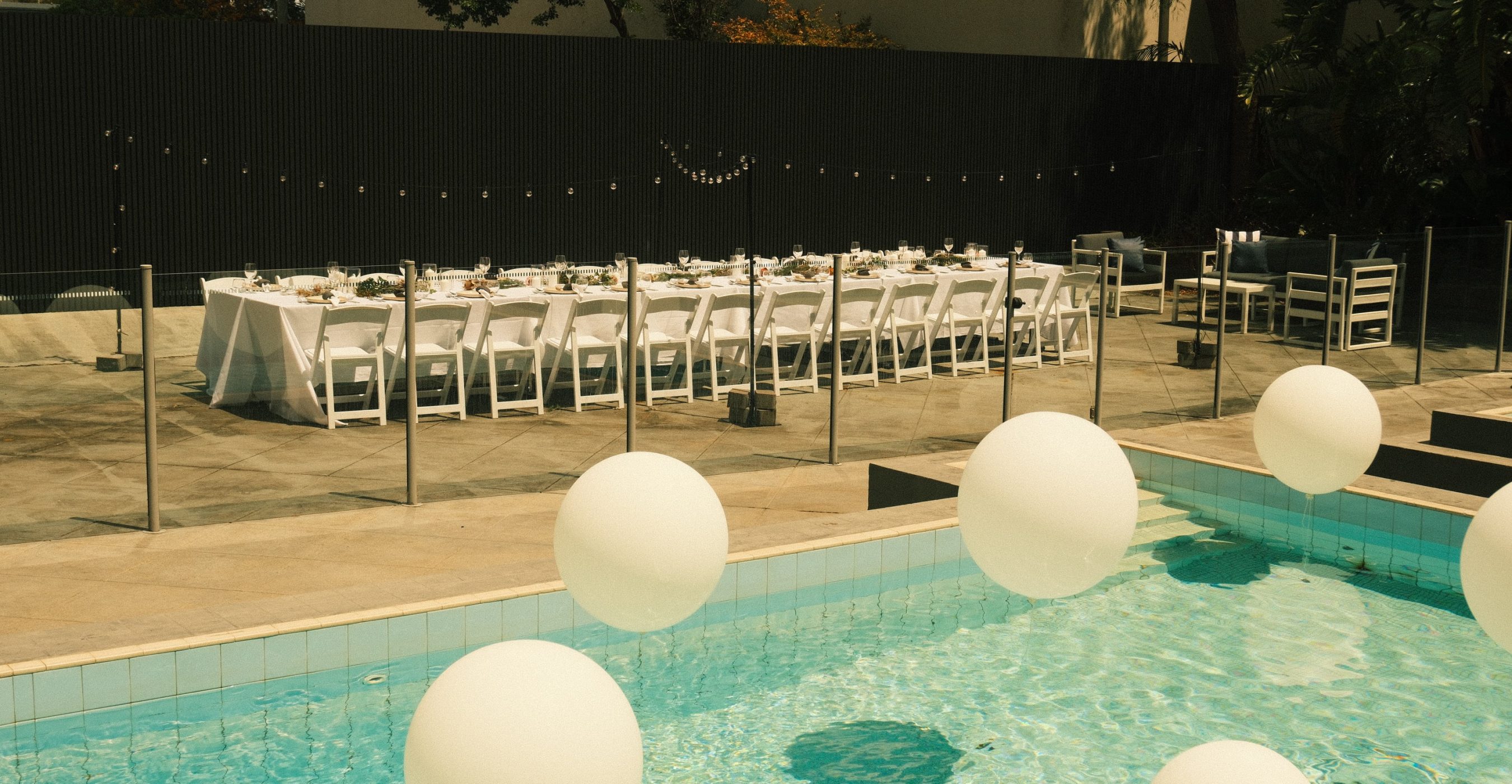 A long banquet table with white chairs sits beside a swimming pool dotted with large floating white spheres.
