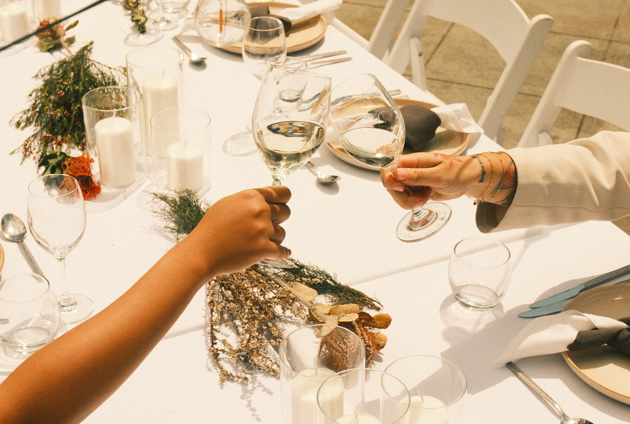 Guests clink wine glasses over a long dining table set with candles, greenery, plates, and white chairs.