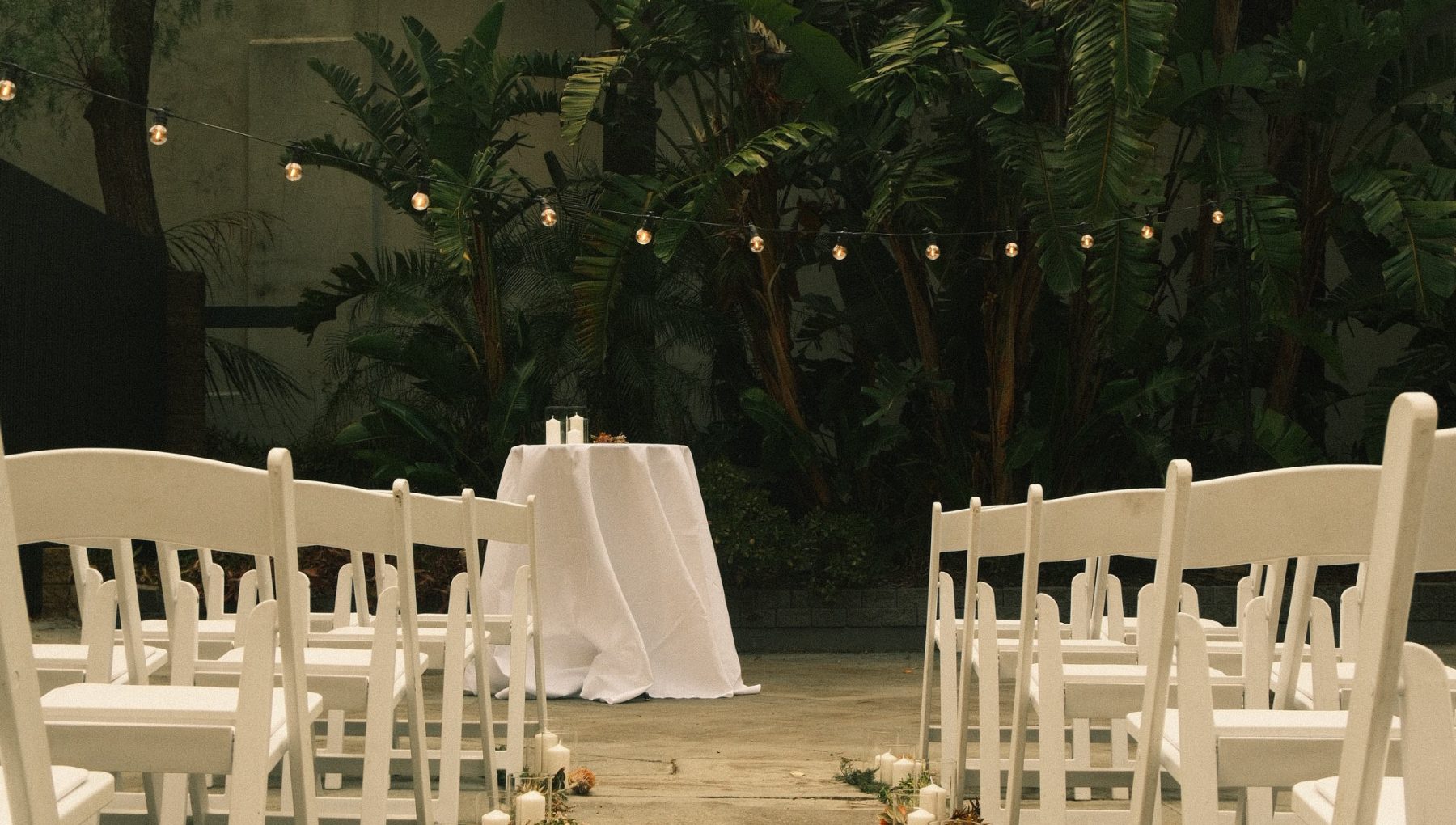 White chairs line an outdoor ceremony aisle leading to a draped table, surrounded by tropical plants and string lights.