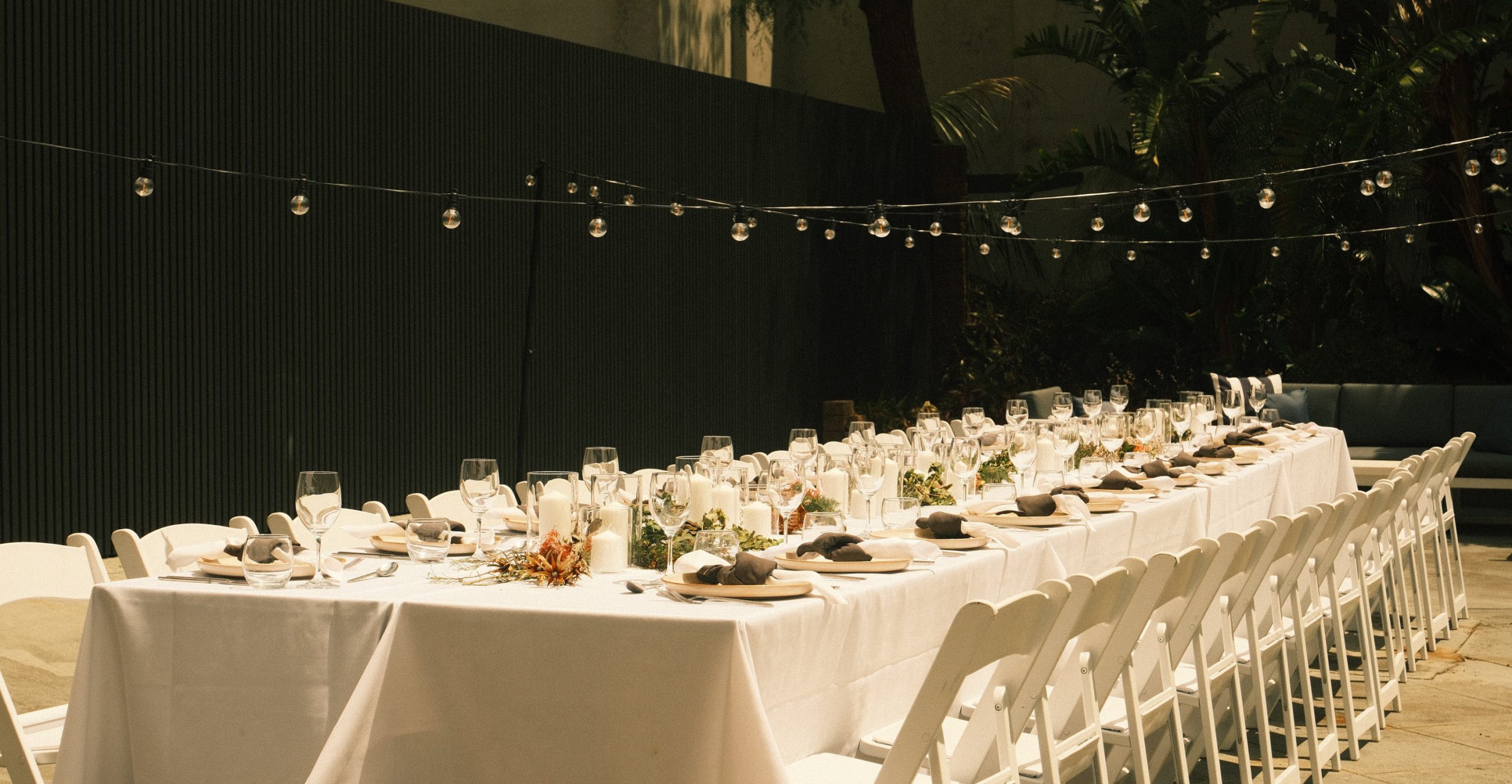 Long white-clothed banquet table set with plates, glassware, candles, and flowers beneath string lights, with white chairs and tropical plants.