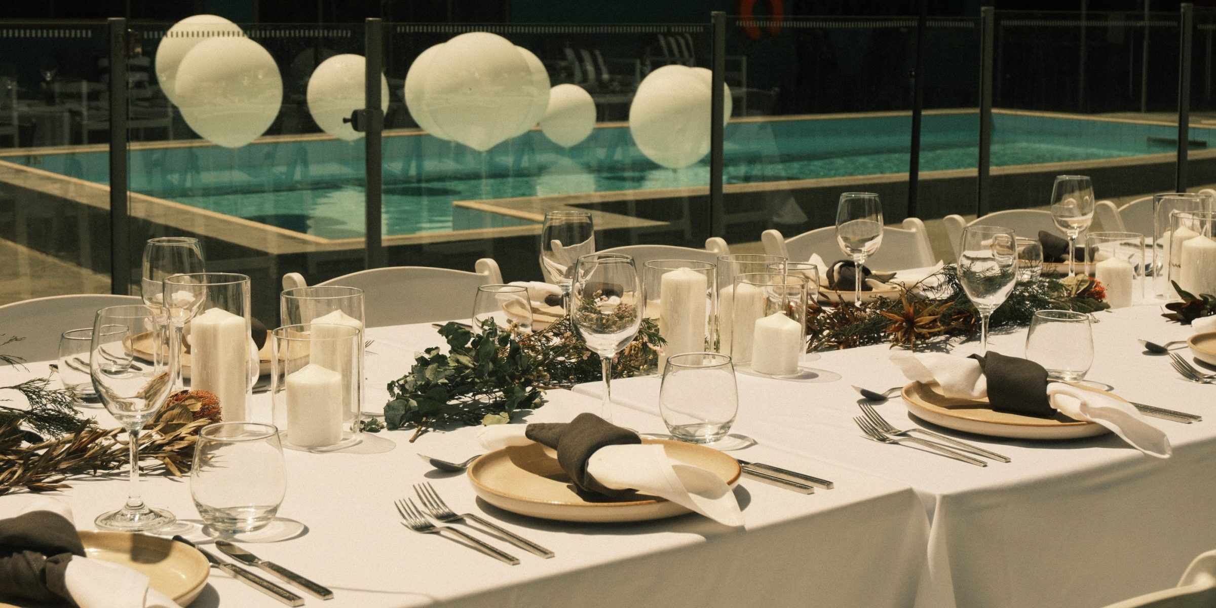 A long banquet table is set with plates, glasses, candles, and greenery beside a turquoise pool and glowing spheres.