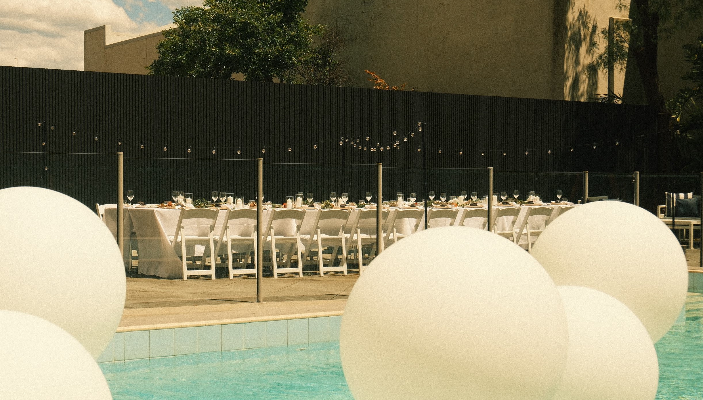 A long outdoor dining table with white chairs sits beside a turquoise pool, framed by large floating white spheres.