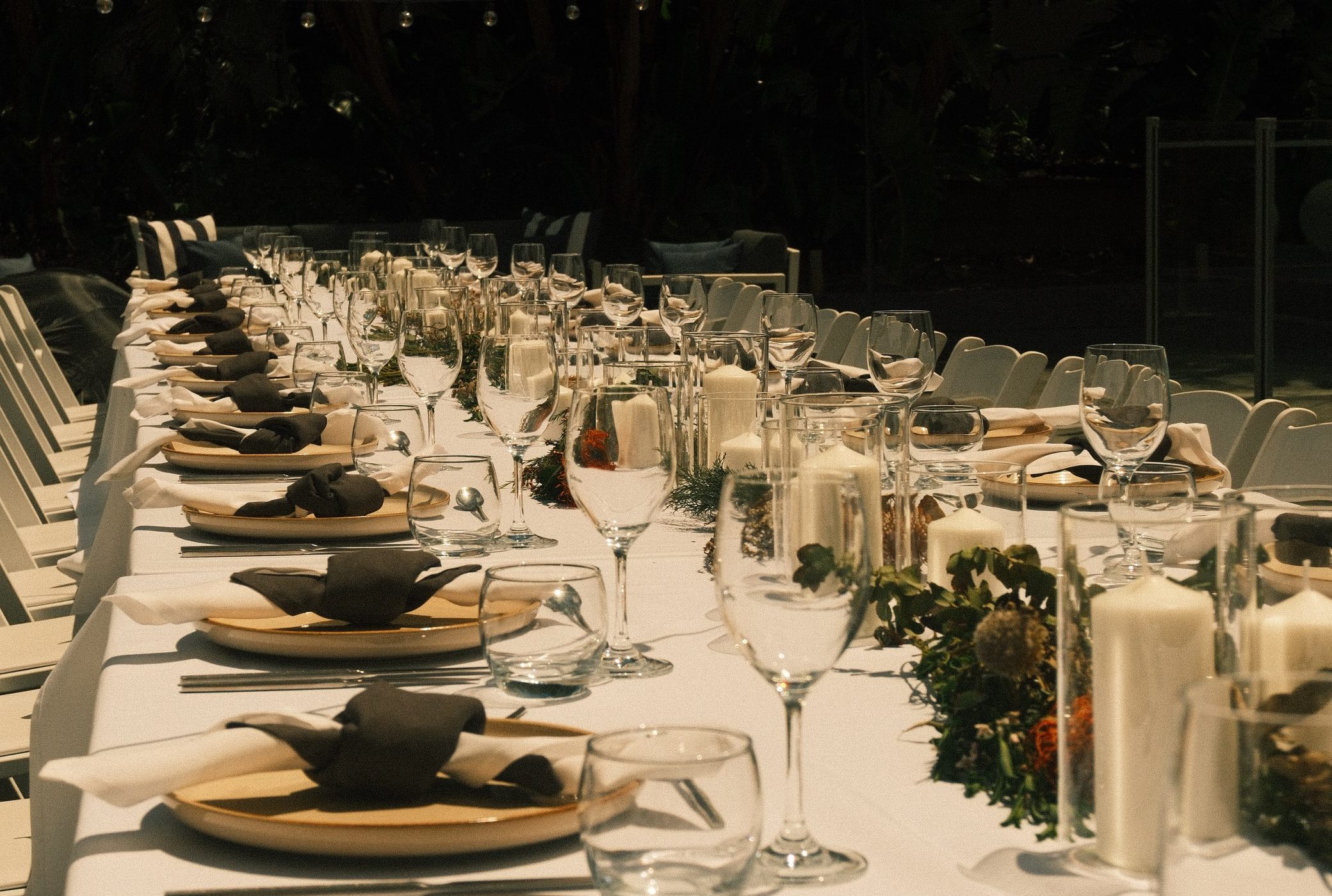 A long dining table set with plates, black napkins, glassware, and greenery beneath string lights among dense foliage.